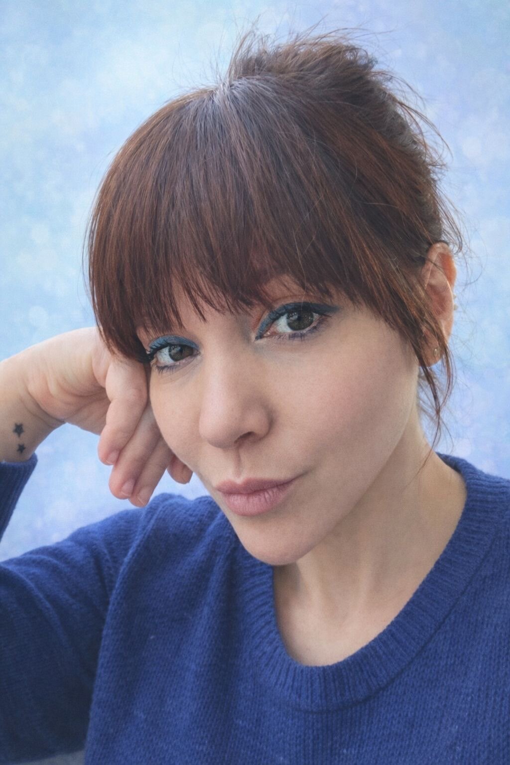 Une femme aux cheveux courts et roux, portant un sweat bleu, pose en regardant l'appareil photo, avec un fond bleu clair.