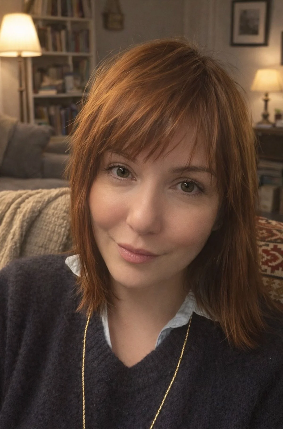 Une femme aux cheveux roux pose pour une photo dans un salon chaleureusement éclairé, avec des étagères remplies de livres et des lampes allumées en arrière-plan.