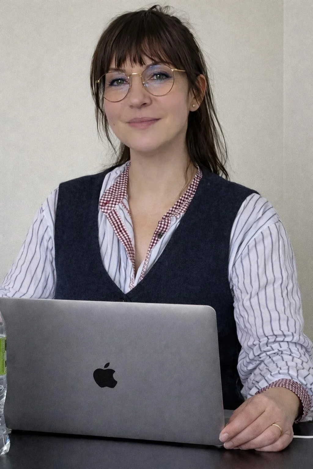 Femme sophistiquée avec lunettes, assise devant un ordinateur portable Apple, portant une chemise à rayures et un gilet, souriant doucement.