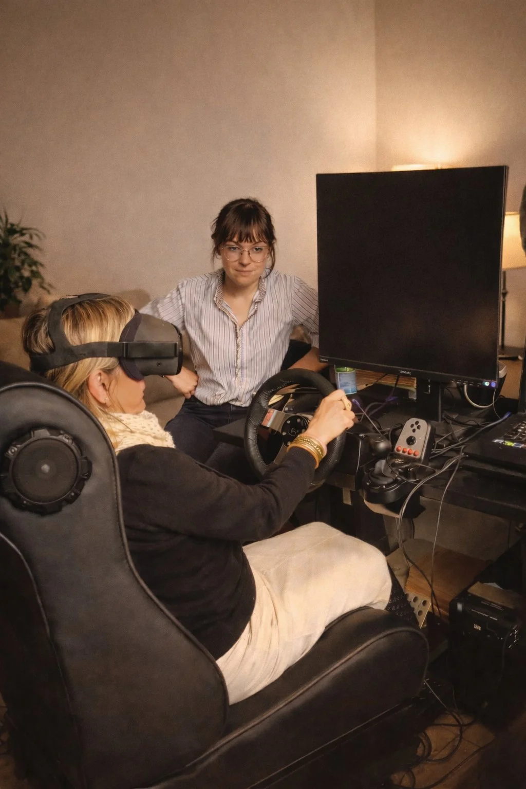 Une femme portant un casque de réalité virtuelle en train de jouer à un jeu vidéo, assise dans un fauteuil, pendant qu'une autre femme observe, dans une pièce avec un ordinateur et un grand écran.