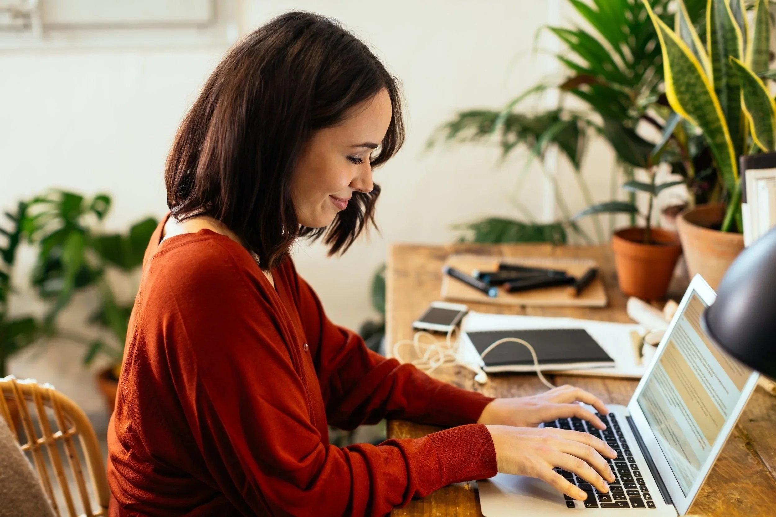 Une femme souriante travaille sur un ordinateur portable dans un espace de travail avec des plantes en pots et des fournitures de bureau.