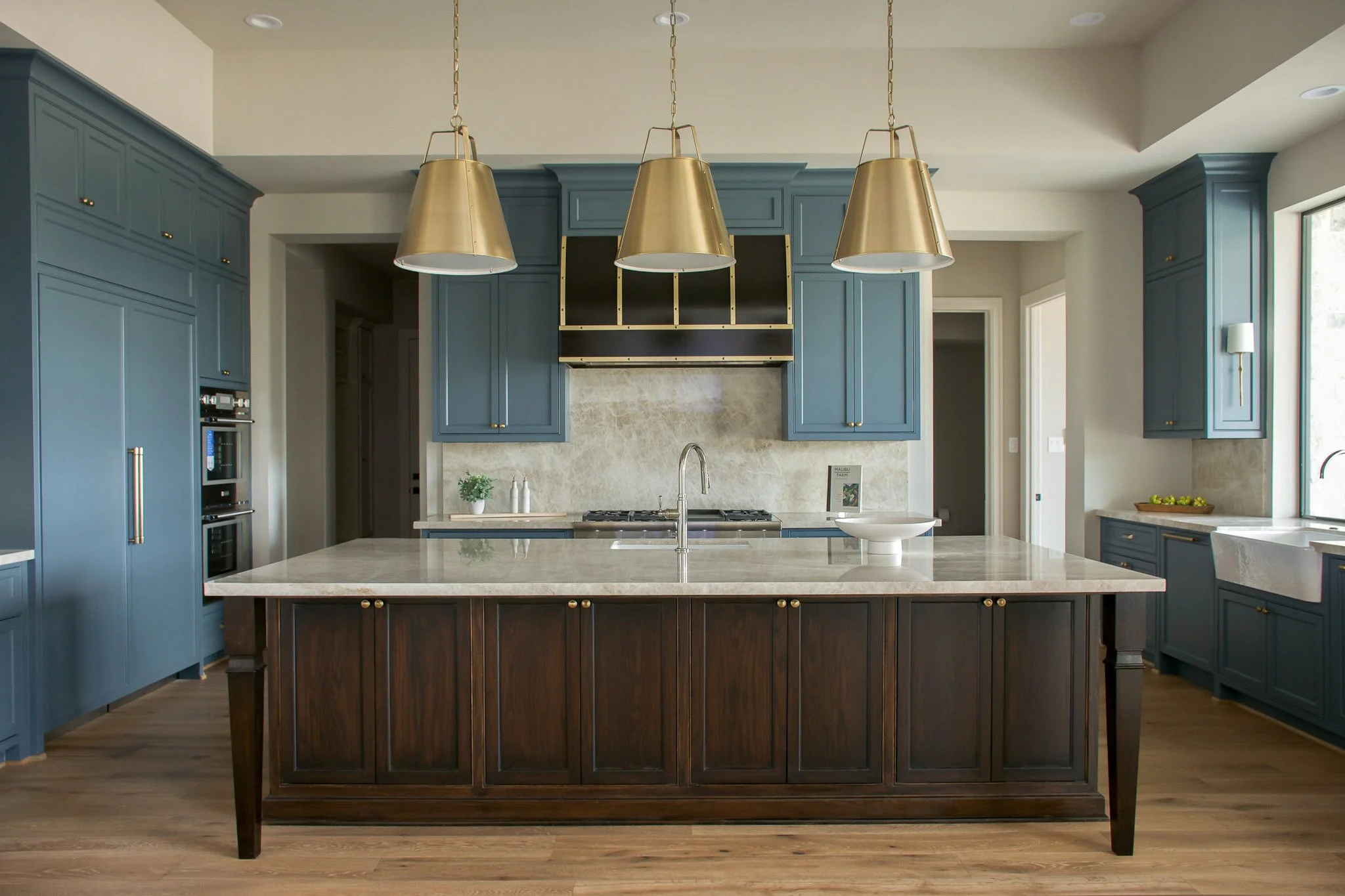 Modern kitchen with blue cabinets, a large island with a dark wood base and light stone countertop, three gold pendant lights, and stainless steel appliances. There's a window to the right and hardwood floors.