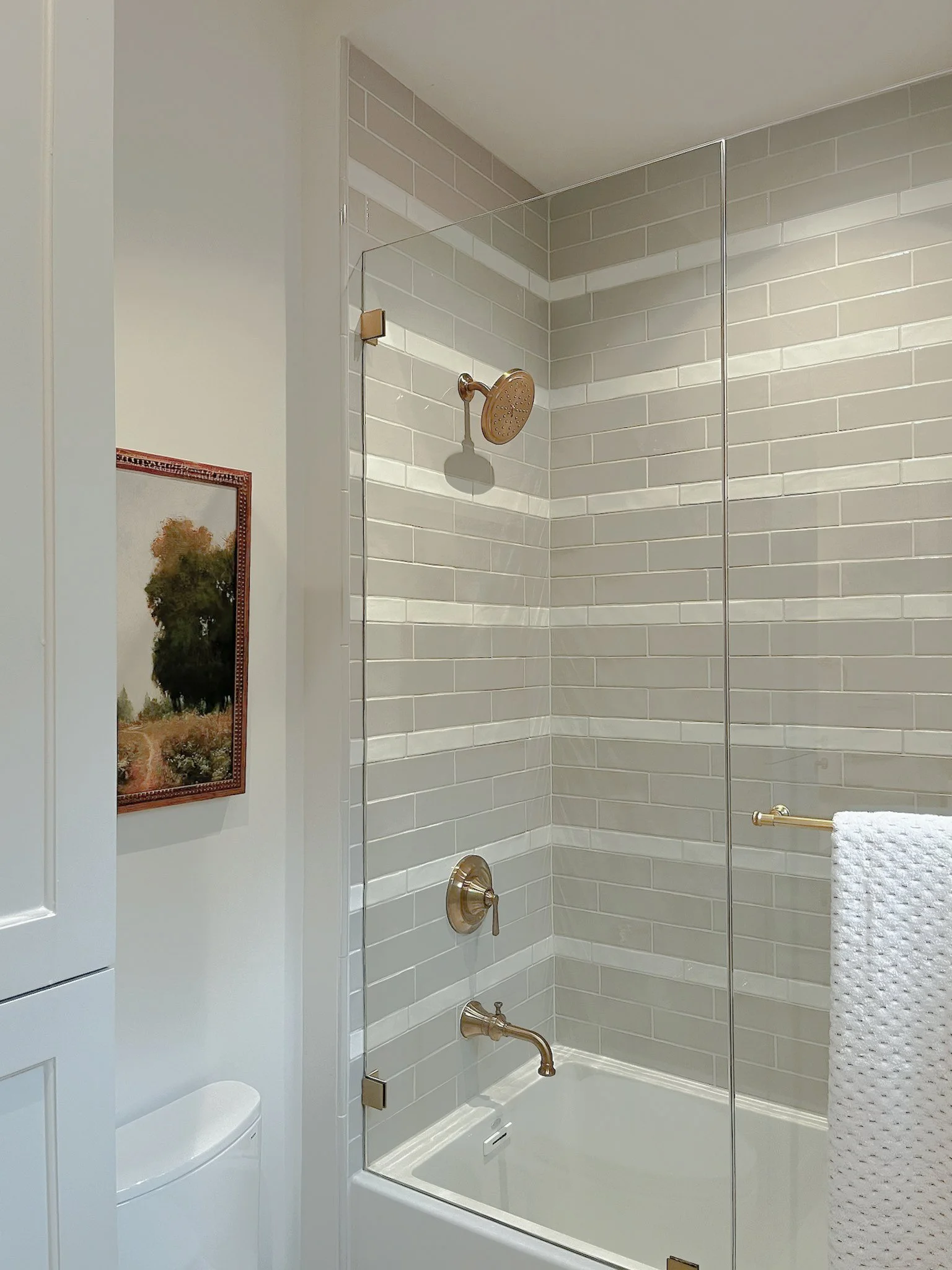 Bathroom shower with beige subway tile walls, a brushed gold showerhead, a towel rack with a white towel, a framed picture of a tree on the wall, and a white bathtub.