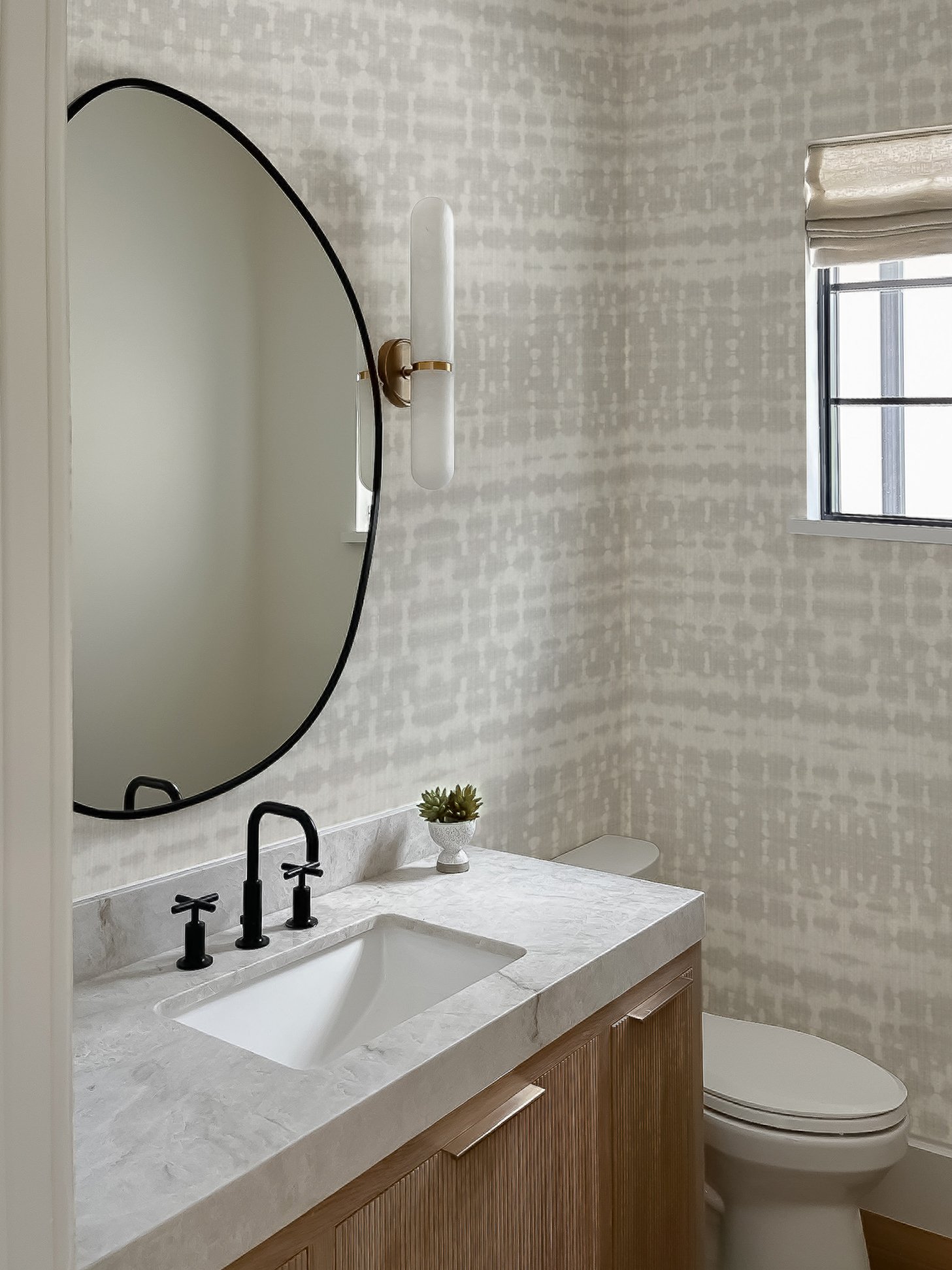Bathroom with a white marble countertop, a undermount sink with black faucet handles, a round mirror with a black frame, a wall-mounted light fixture, a small potted plant, a window with a beige shade, and a toilet.