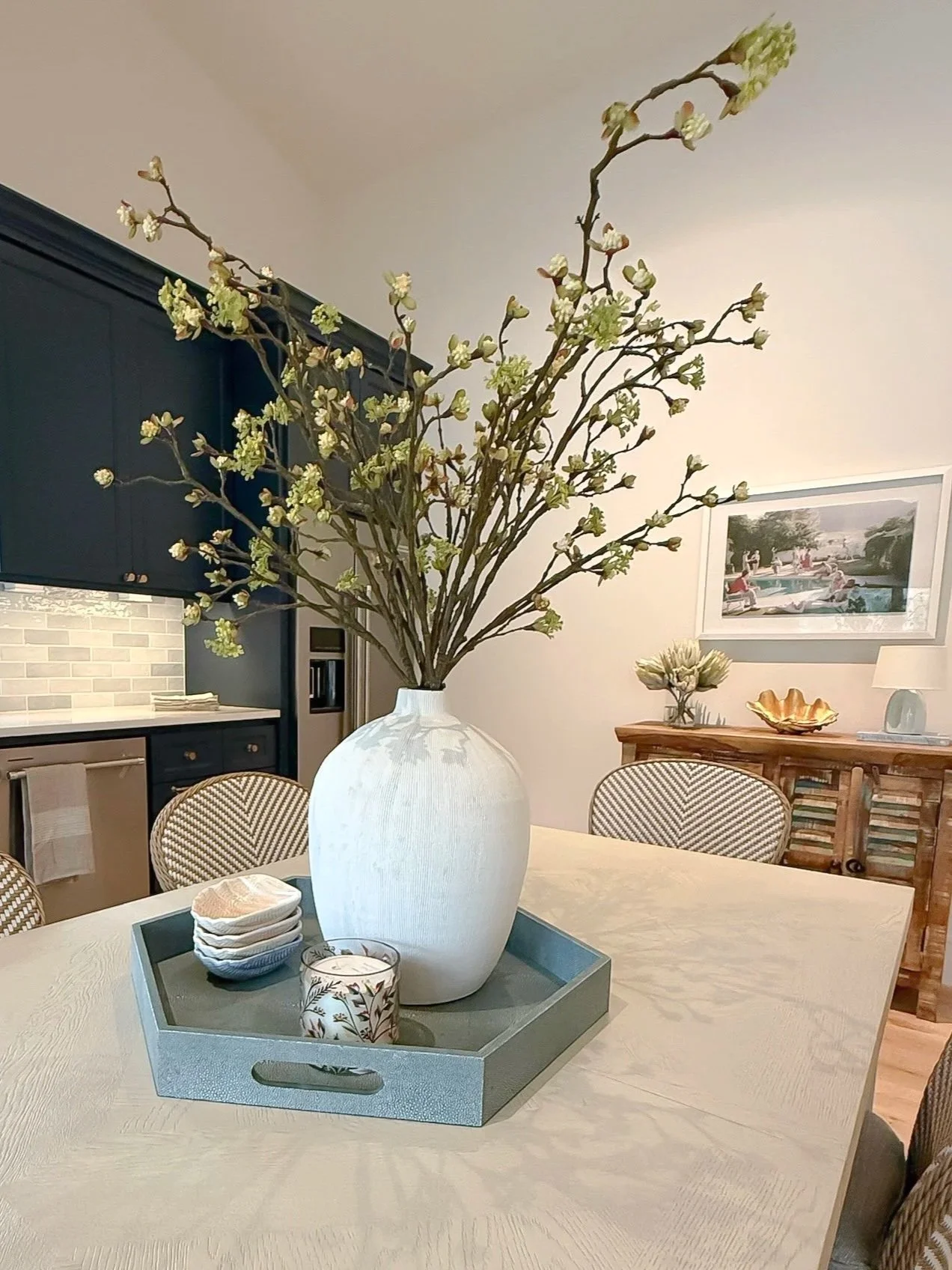 A large white vase with a branch of blooming flowers and green leaves placed on a dining table. In the background, there is a kitchen and a sideboard with decorative items and a framed artwork on the wall.