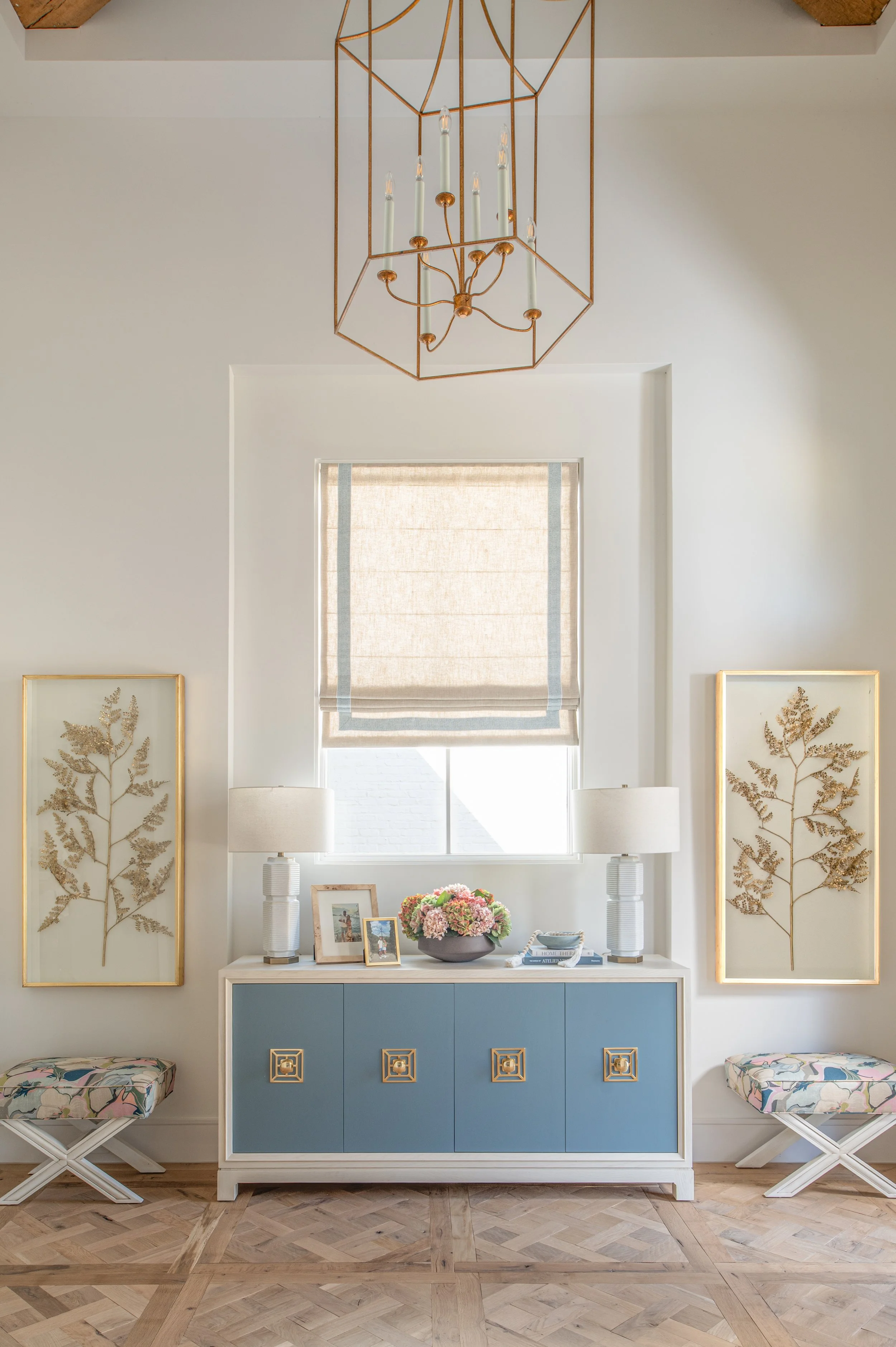 Interior view of a living room with a blue and white sideboard, framed botanical artwork, a window with a beige shade, a symmetrical pair of white lamps, and decorative items including framed photos and a flower arrangement. A chandelier hangs from t