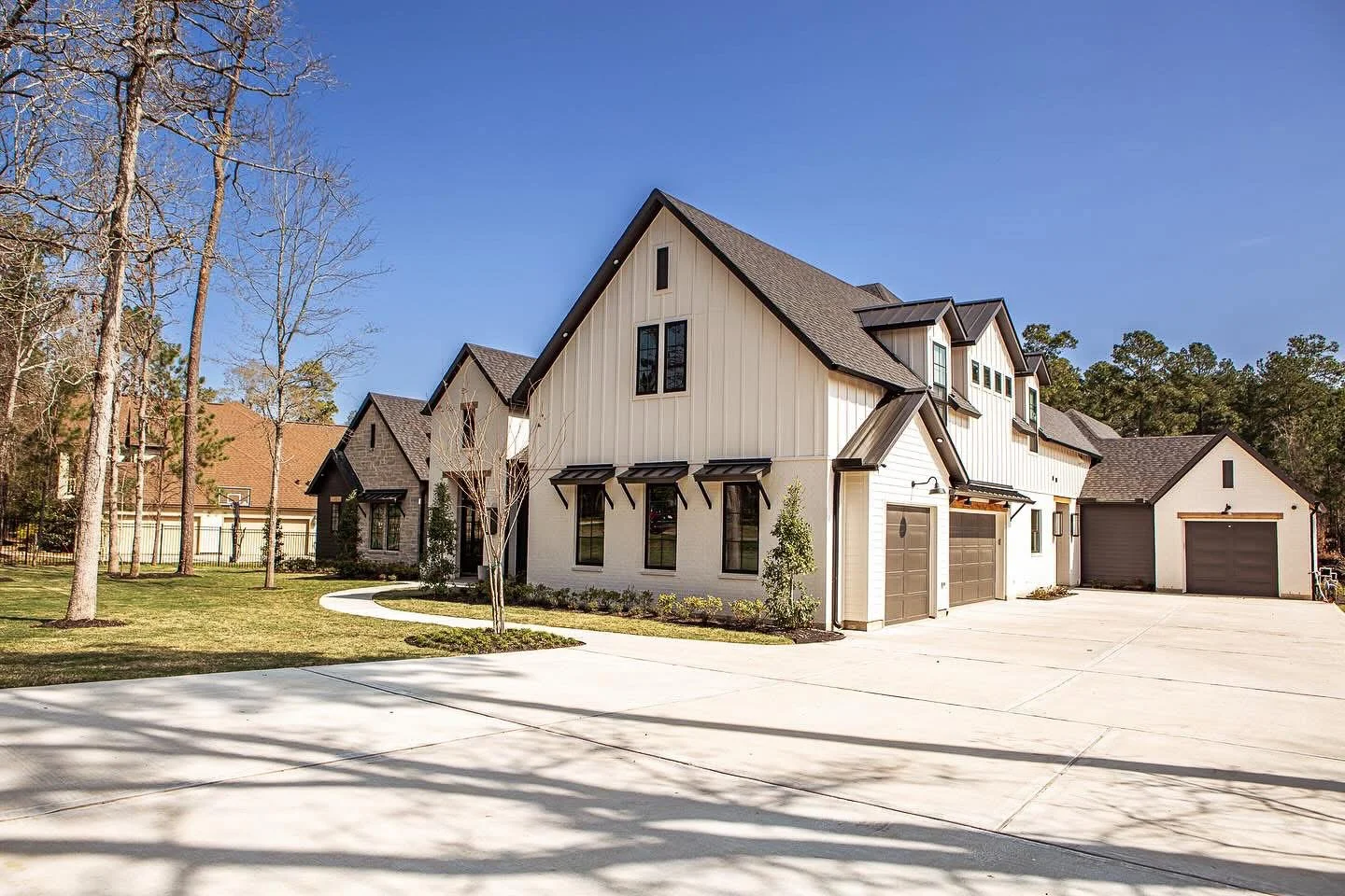 A modern two-story house with a spacious driveway, surrounded by trees, featuring a mix of horizontal and vertical siding, multiple gables, and a three-car garage.