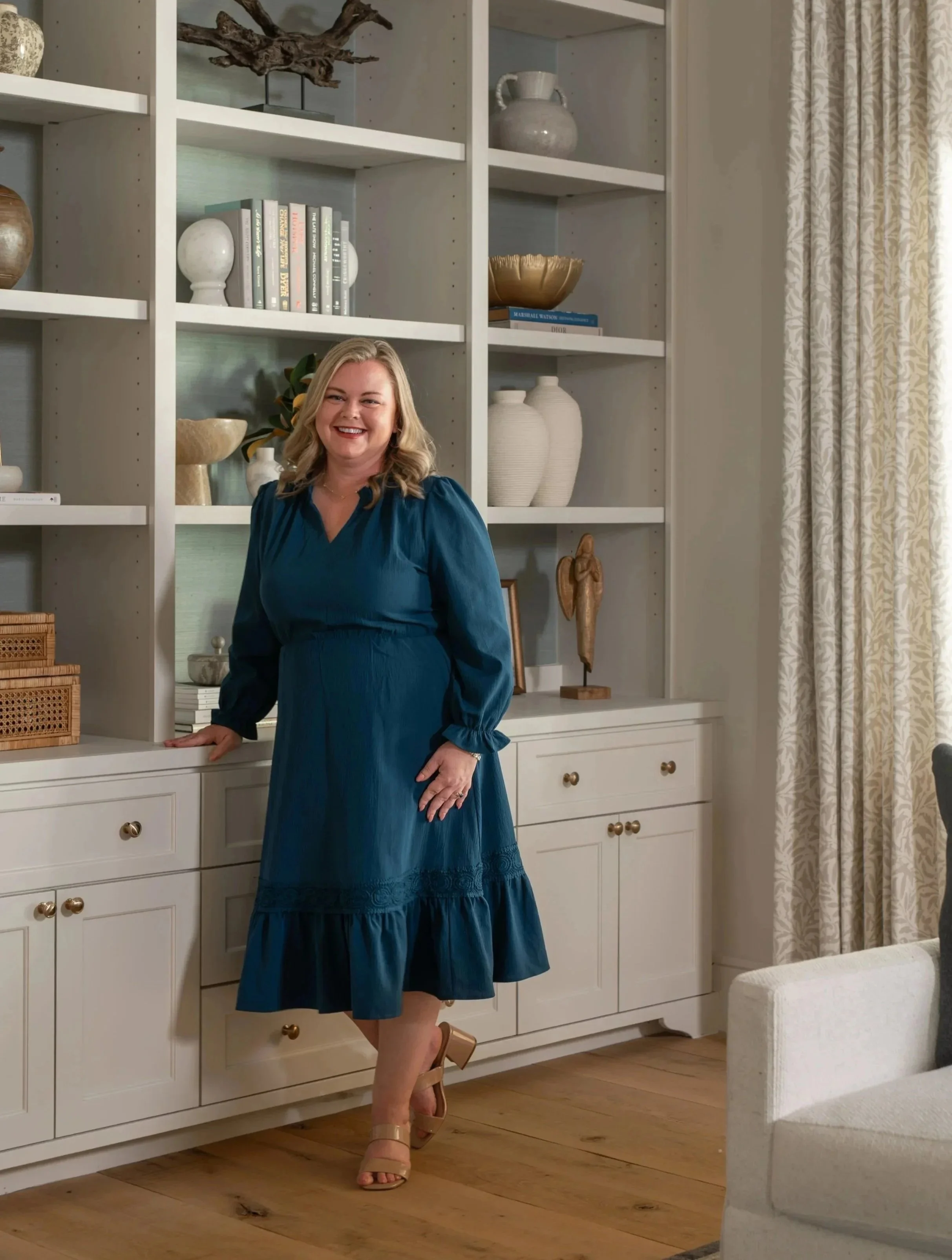 A woman smiling and standing in a room with white bookcases filled with decorative items, plants, and books. She is wearing a blue dress and open-toed heels, with one hand resting on a white cabinet and the other on her hip. There is a curtain with a light pattern and a light-colored sofa in the foreground.