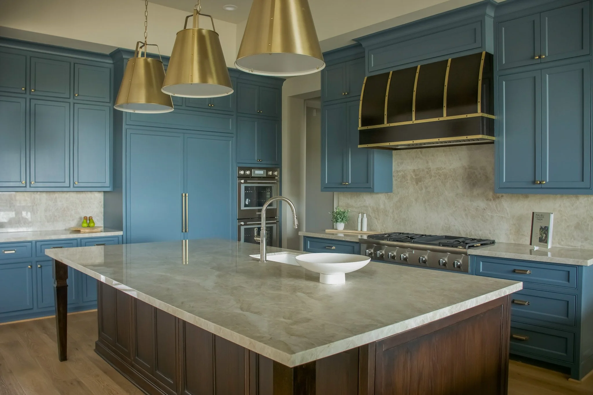 Modern kitchen with blue cabinets, a large island with a beige marble countertop, a stainless steel oven, and three gold pendant lights hanging above the island.