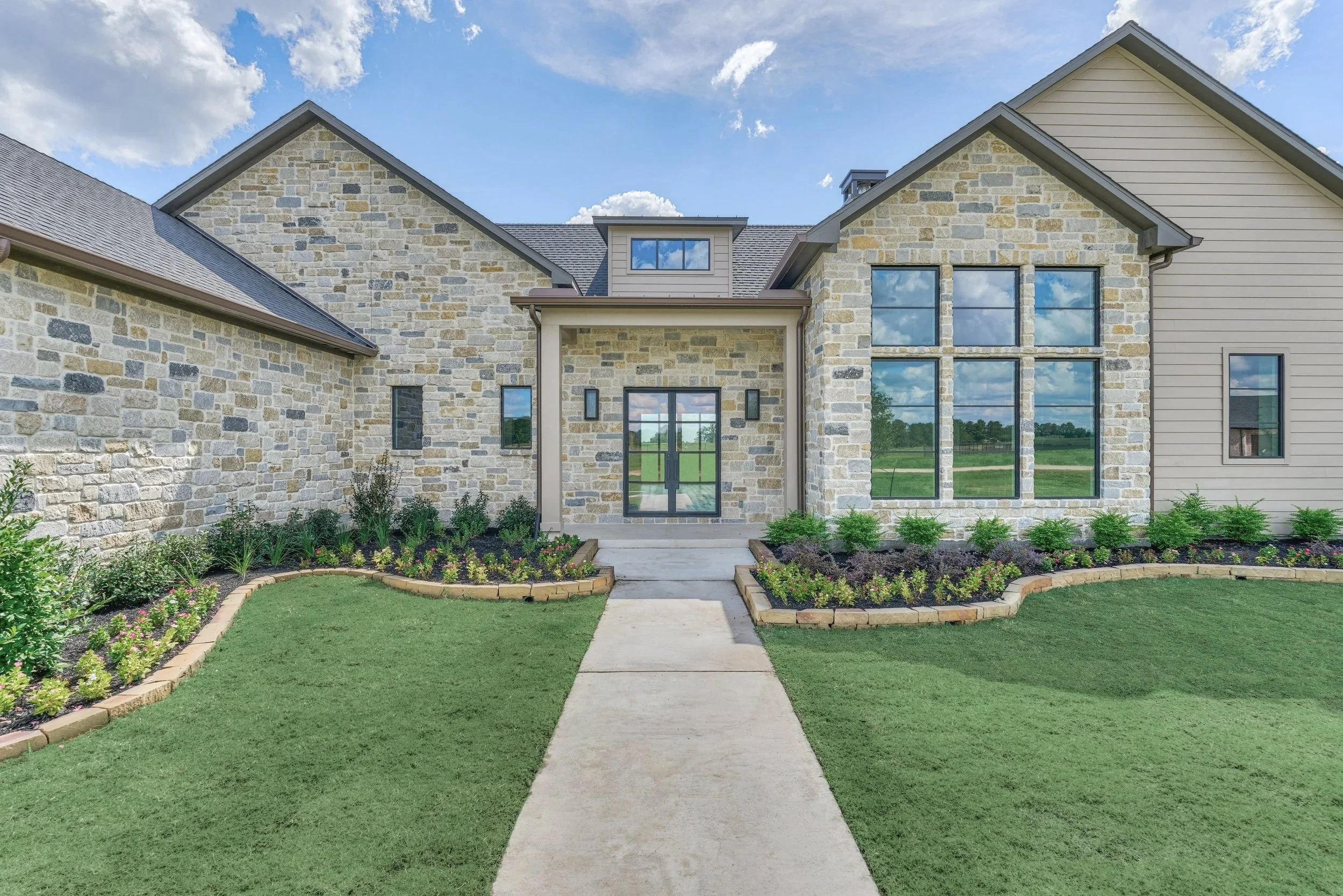 Front view of a modern house with a combination of stone and siding exterior, large windows, a concrete walkway leading to the entrance, and a well-maintained green lawn and flower beds.