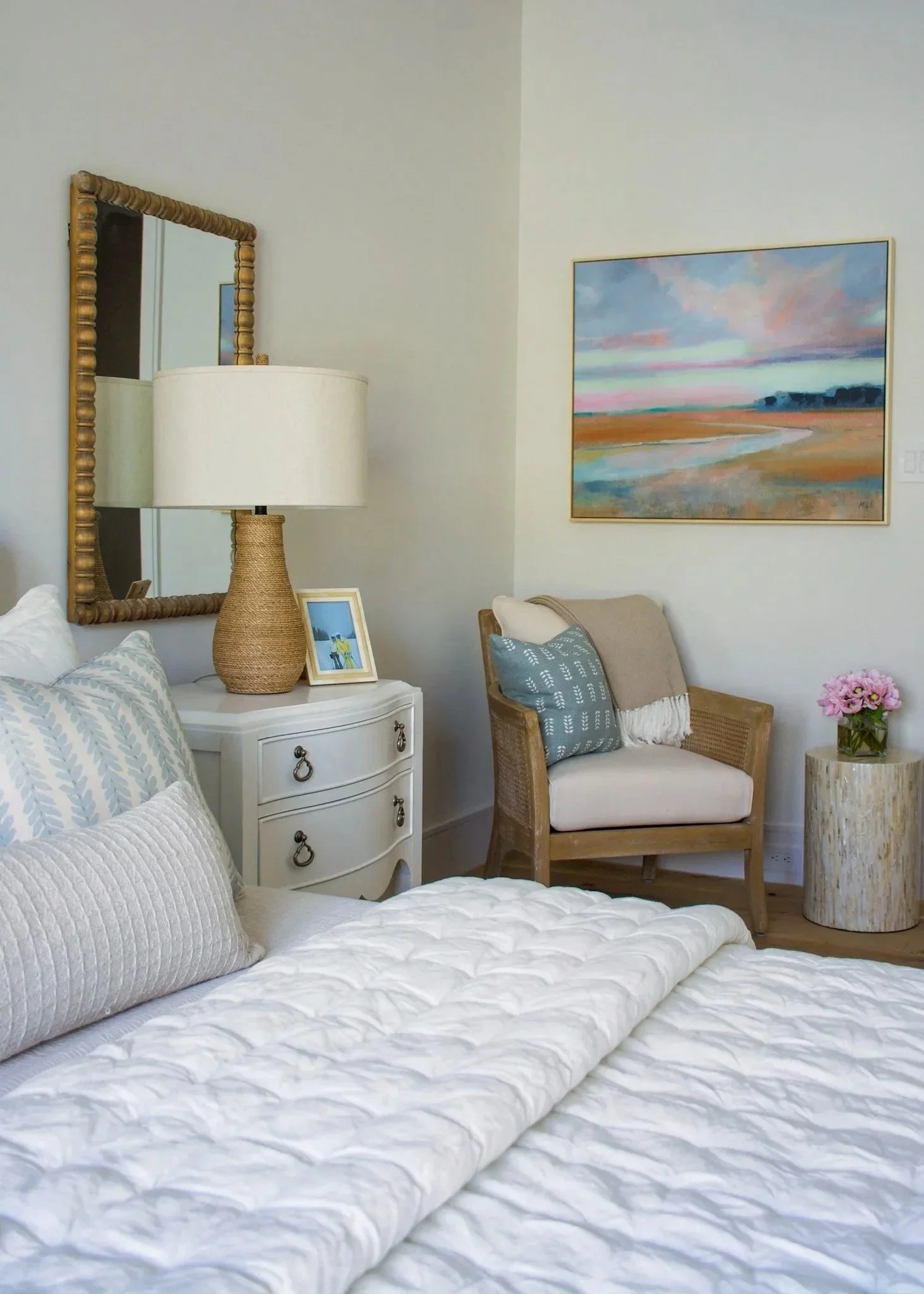 A cozy bedroom with white bedding, a beige rolled-up pillow, a white nightstand with a basket-style lamp, framed photo, and a large mirror. There's a wooden armchair with pillows, a side table with pink flowers, and a landscape painting on the wall.