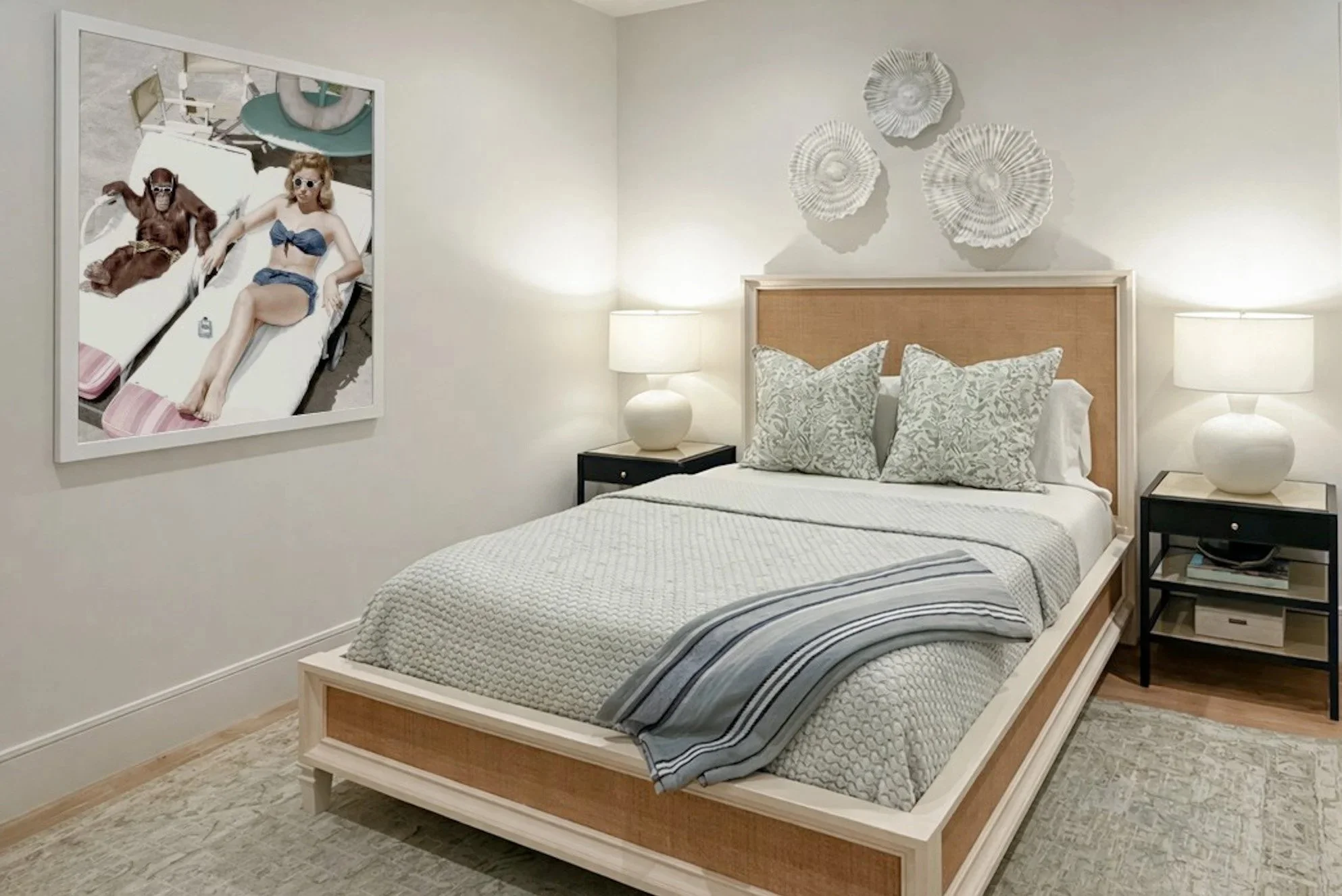 A bedroom with a wooden bed frame, white patterned bedding, and pillows, two white lamps on nightstands, wall decorations resembling seashells or fan-shaped objects, and a large framed photo on the wall depicting a woman and a chimpanzee relaxing on 