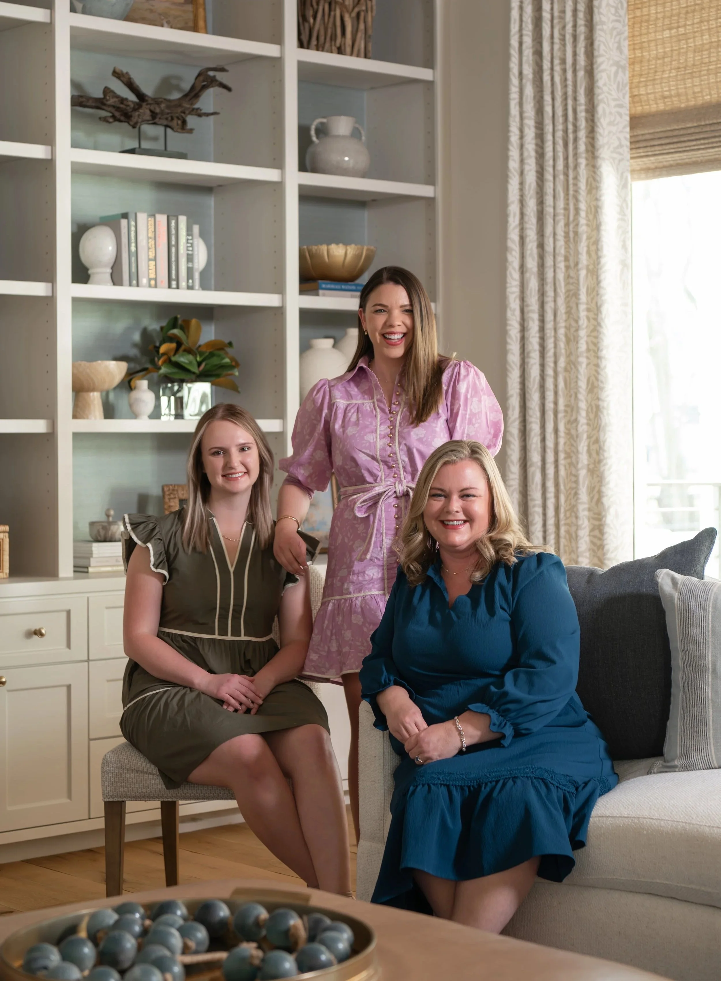 Four women sitting and standing in a living room with a bookshelf and window in the background. One woman in a pink dress standing behind two seated women, one in a gray dress and the other in a blue dress.