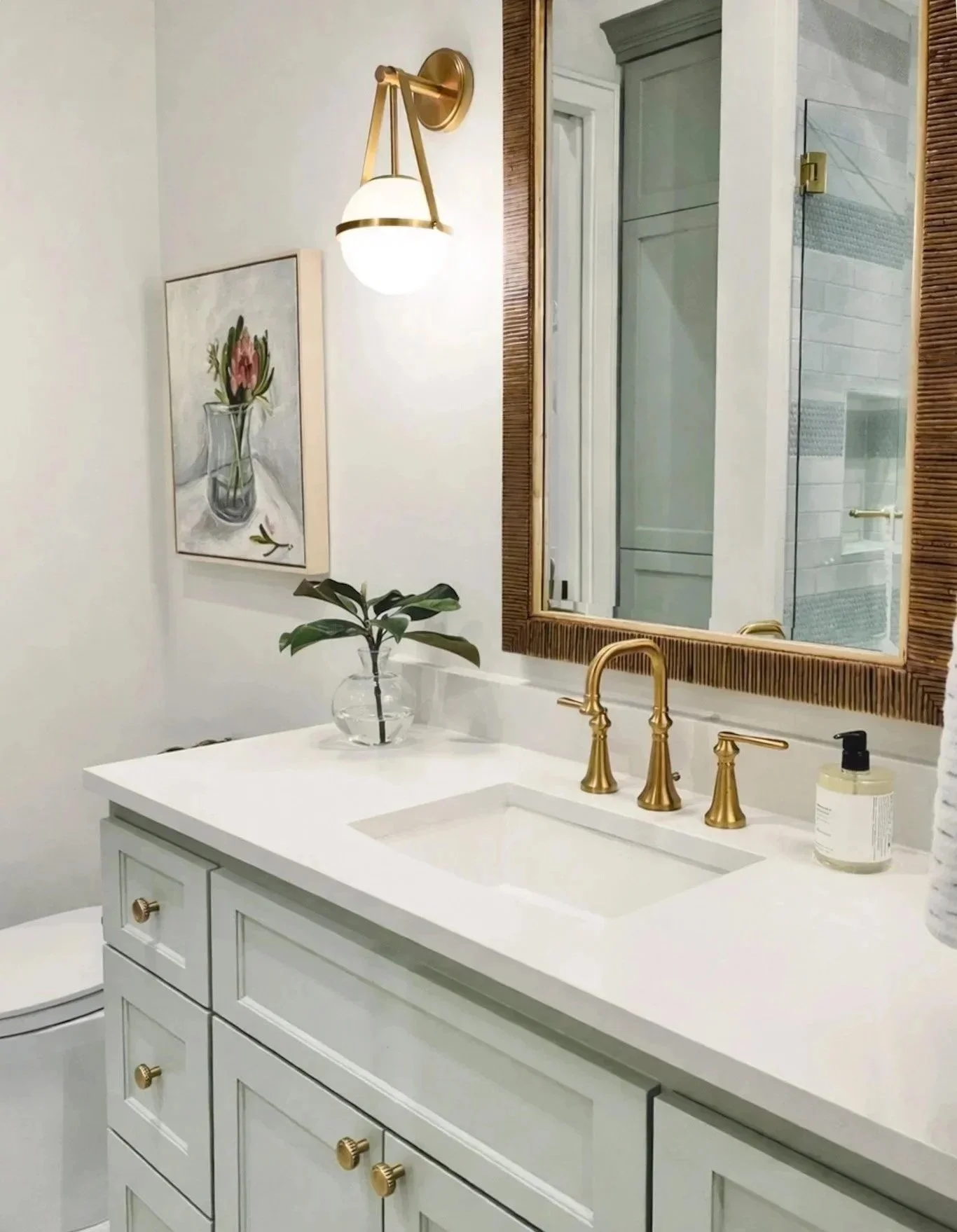 Bathroom vanity with white cabinets, gold fixtures, a large mirror with a wooden frame, a vessel sink with a gold faucet, a bottle of soap, a glass vase with a green plant, and wall art of a pink flower in a glass vase.