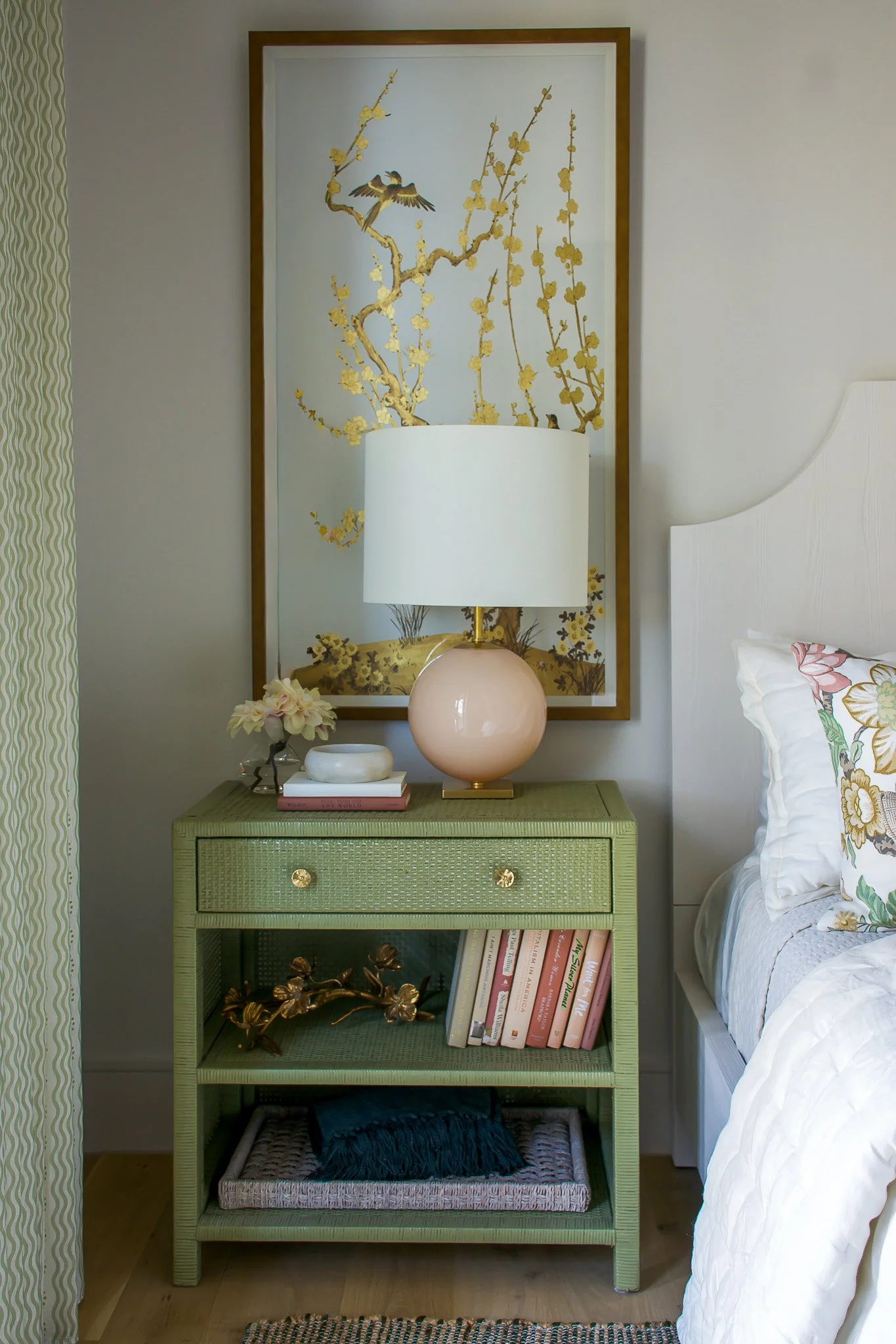 A bedroom with a green nightstand, a white lamp, a bowl, pink and white books, and a small flower vase. Behind the lamp is a large framed artwork of a golden flowering tree with birds. To the right, part of a white bed with floral pillows is visible.
