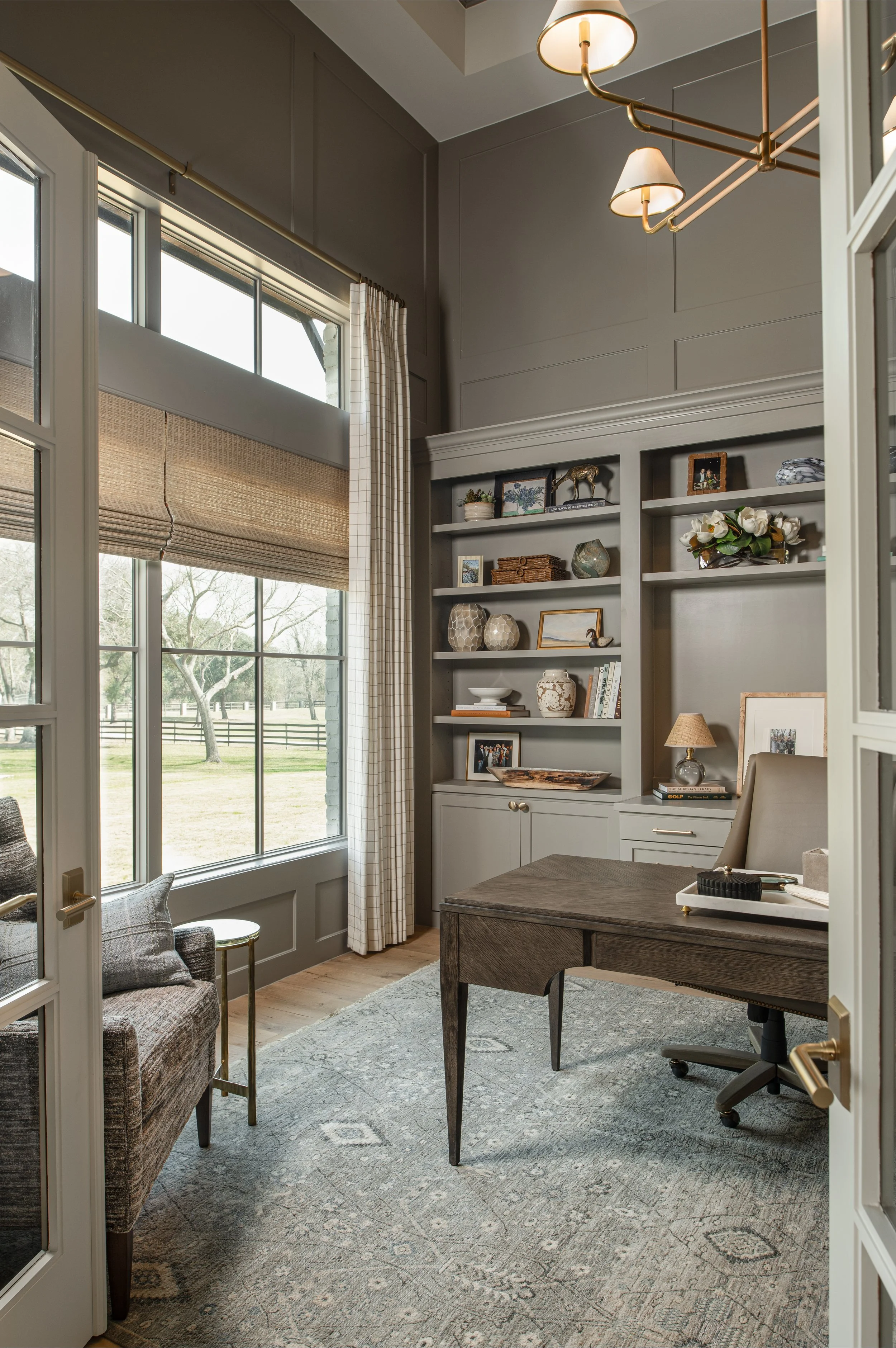 A home office with large windows, a built-in gray bookshelf filled with decorative items, and a desk with a chair, a lamp, and a tray. The room has a gray patterned rug, and the windows have light curtains and woven shades. Outside, there is a green 