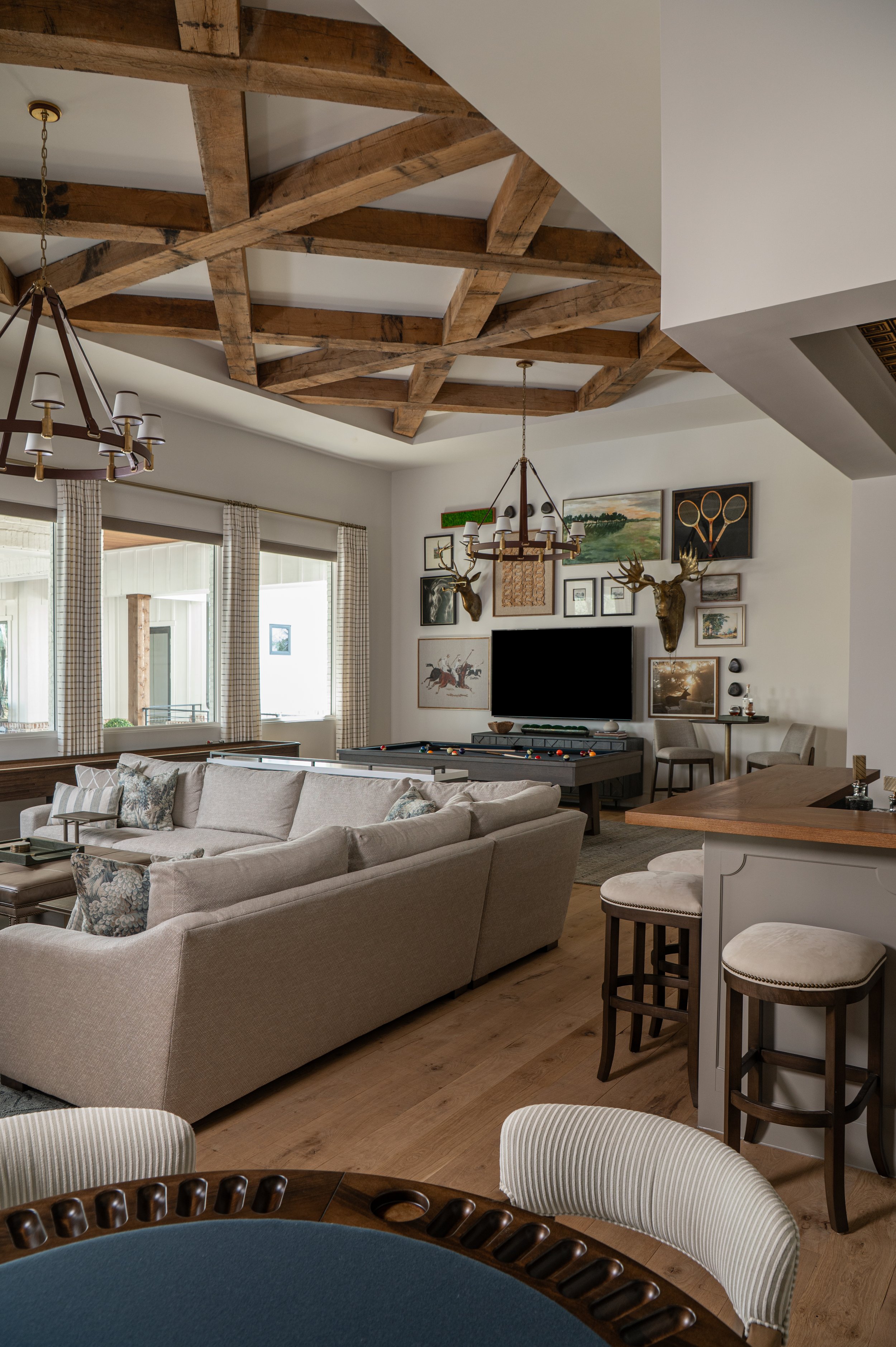 Living room with beige sofa, wooden dining table, patterned curtains, wall art including paintings and taxidermy deer heads, pool table, and wood beam ceiling.