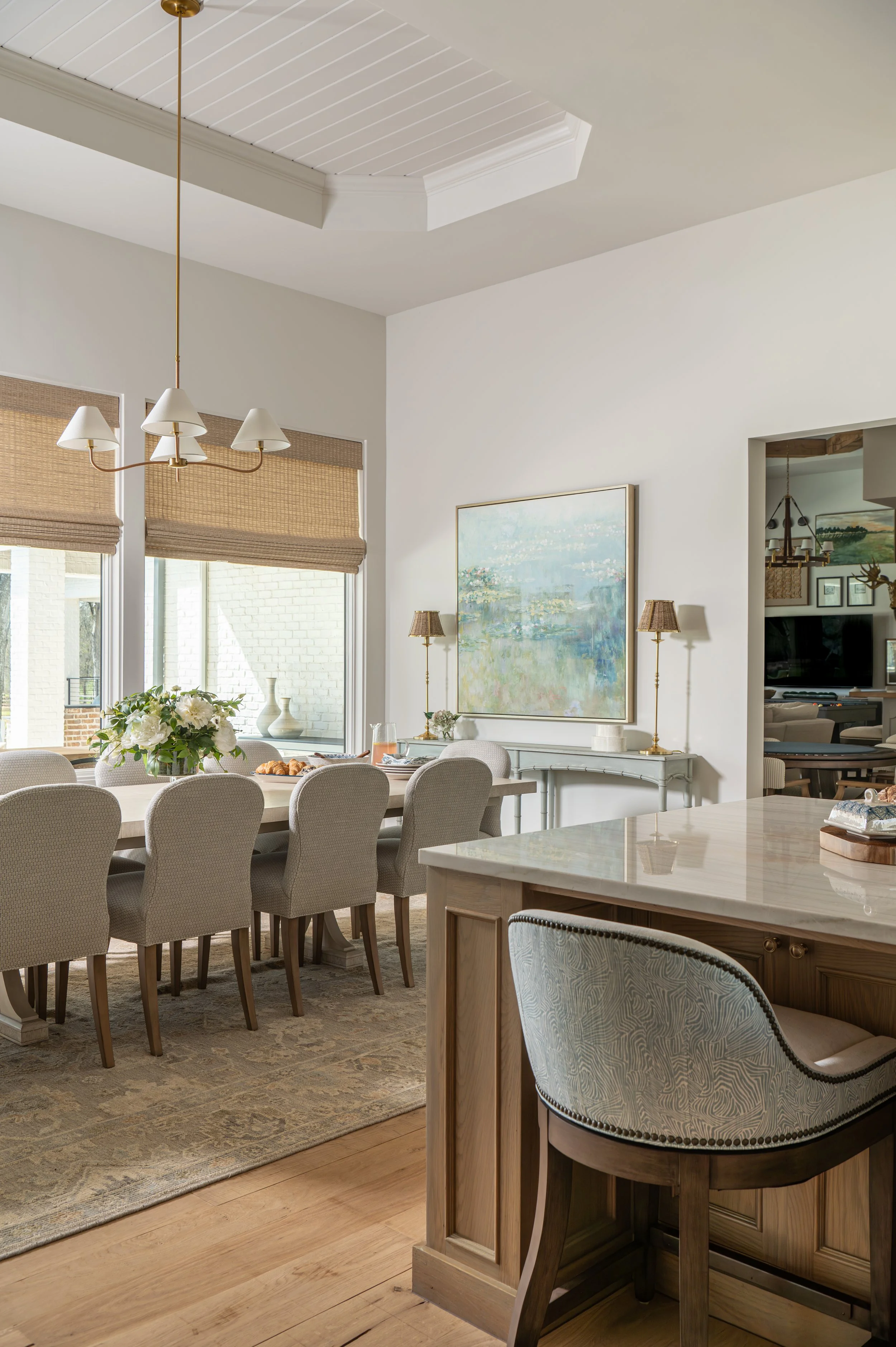 Interior of a dining area with a large table surrounded by beige upholstered chairs, a chandelier, large windows with woven shades, artwork on the wall, and a view into a living room area with additional seating and decor.