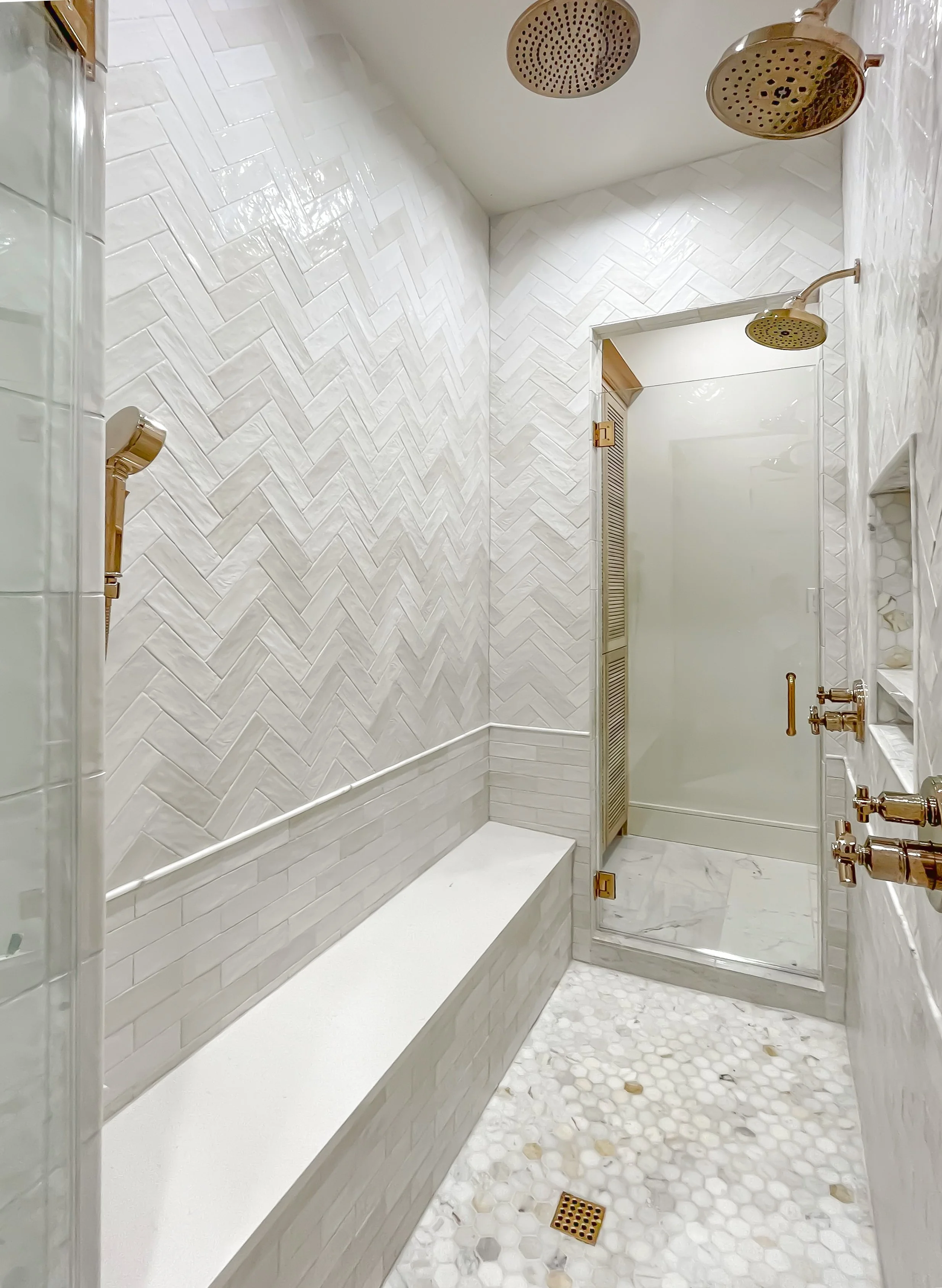 Modern shower with dual rainfall shower heads, white herringbone tile walls, marble flooring, glass enclosure, and built-in bench.