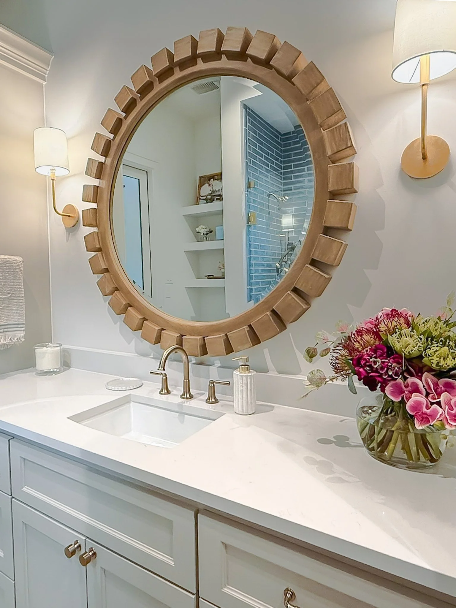 A white bathroom vanity with a white countertop, a gold faucet, a small soap dispenser, a candle, a bouquet of pink and white flowers in a glass vase, a large round mirror with a wooden frame, two wall-mounted light fixtures, a shower with blue brick