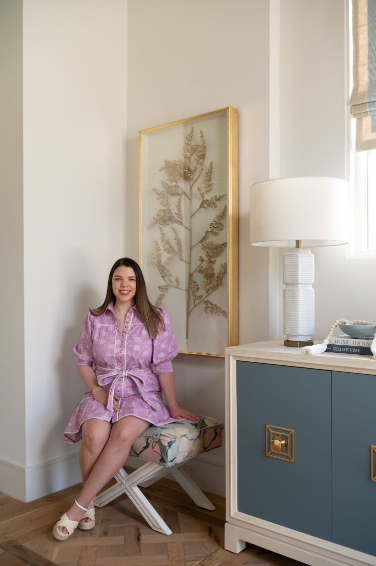 A woman in a pink dress sitting on a floral bench in a room with a blue cabinet, a large white lamp, and a framed art piece with a gold leaf design.