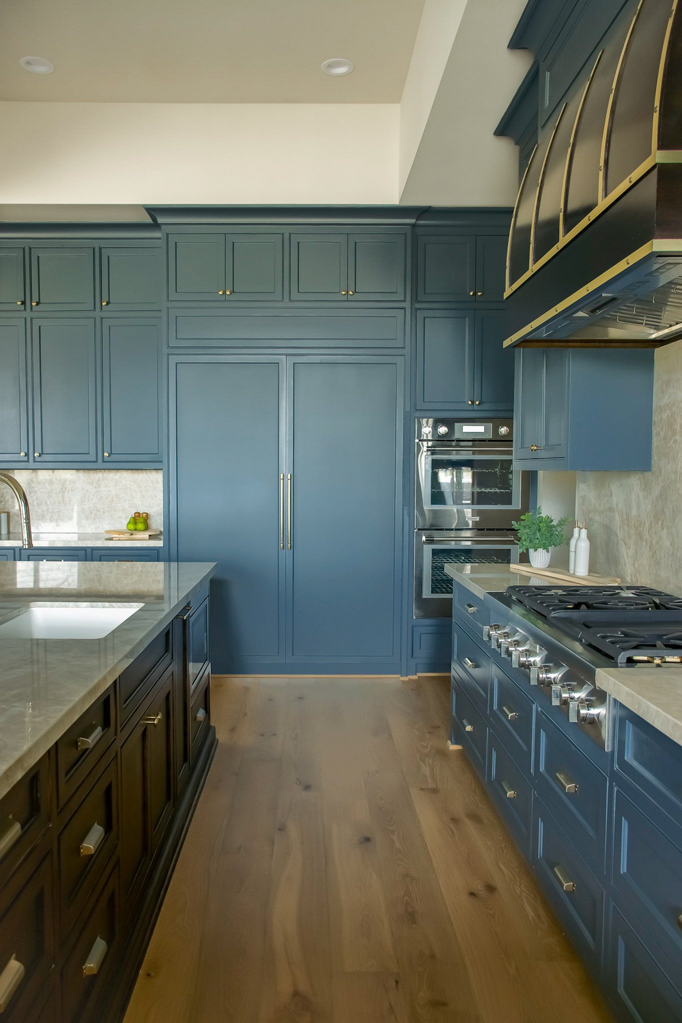 A modern kitchen with dark blue cabinets, a stainless steel oven, and a beige marble island. The kitchen features light wood flooring and decorative vases on a small table.