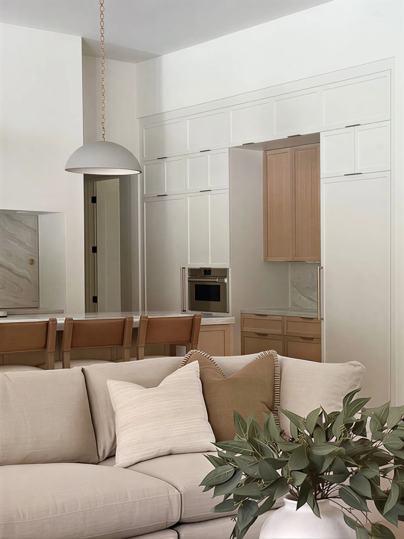 Interior of a modern, minimalistic living room with a beige sectional sofa, decorative pillows, a potted plant, and a kitchen area with white cabinetry, a wooden door, and a hanging pendant lamp.