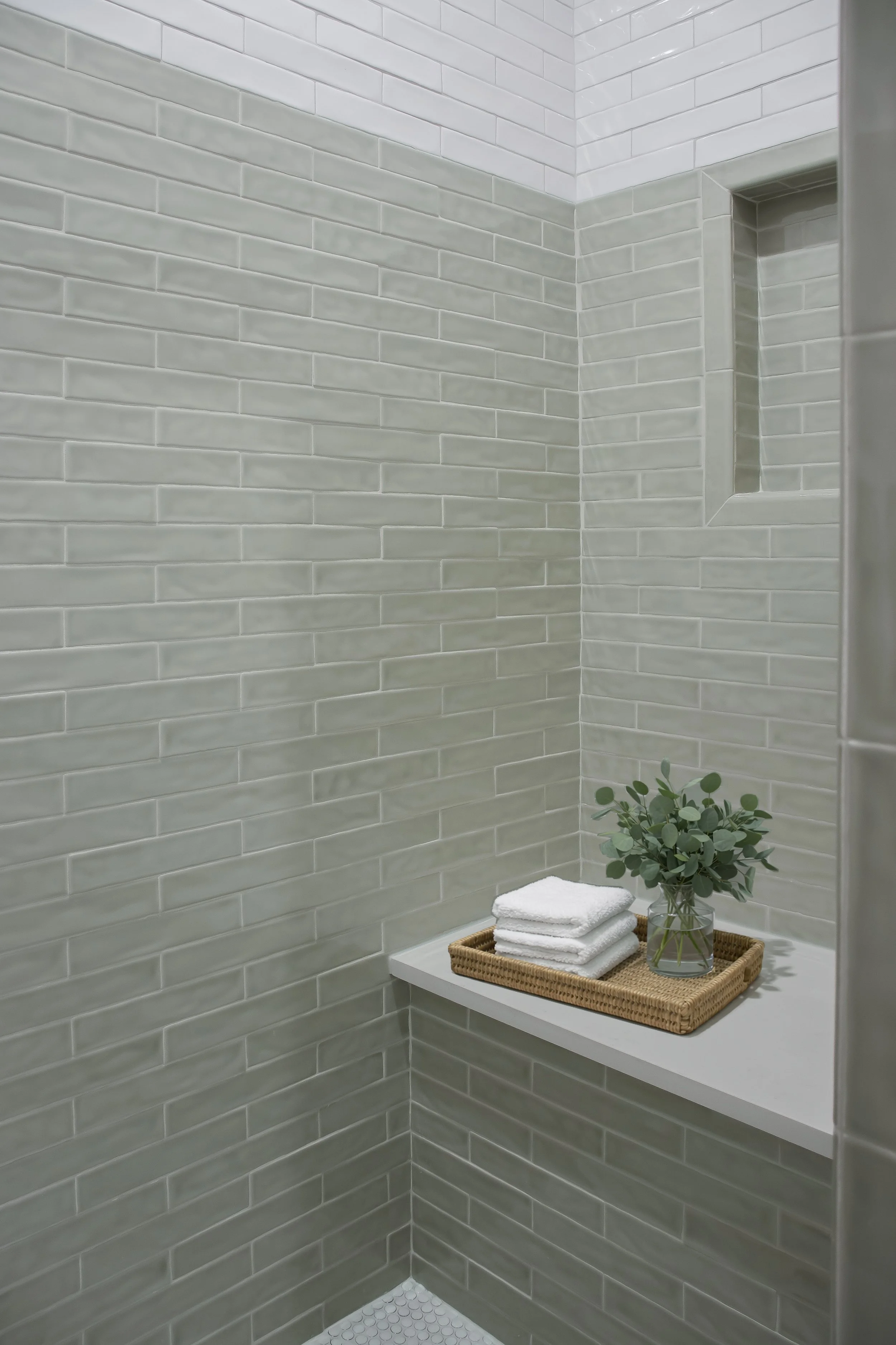 A corner of a modern bathroom with light gray subway tile walls, a small built-in niche in the wall, and a white floating shelf holding a tray with folded white towels and a glass vase with green leaves.