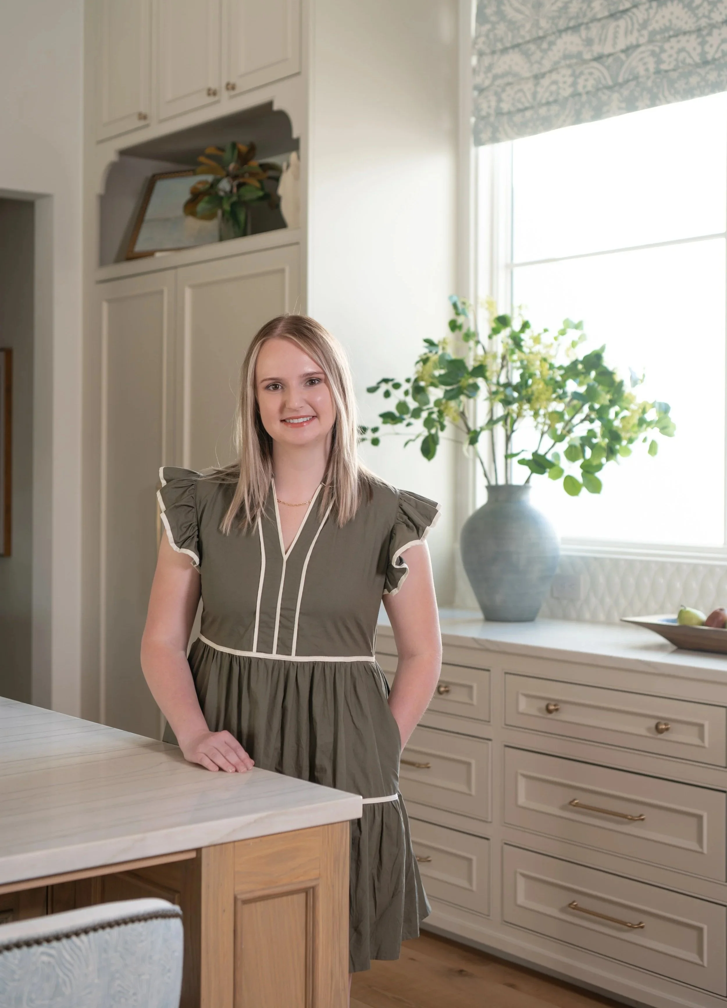 A woman with long blonde hair standing in a kitchen, smiling, with beige cabinetry, white countertops, and a potted plant near a large window with patterned window treatments.