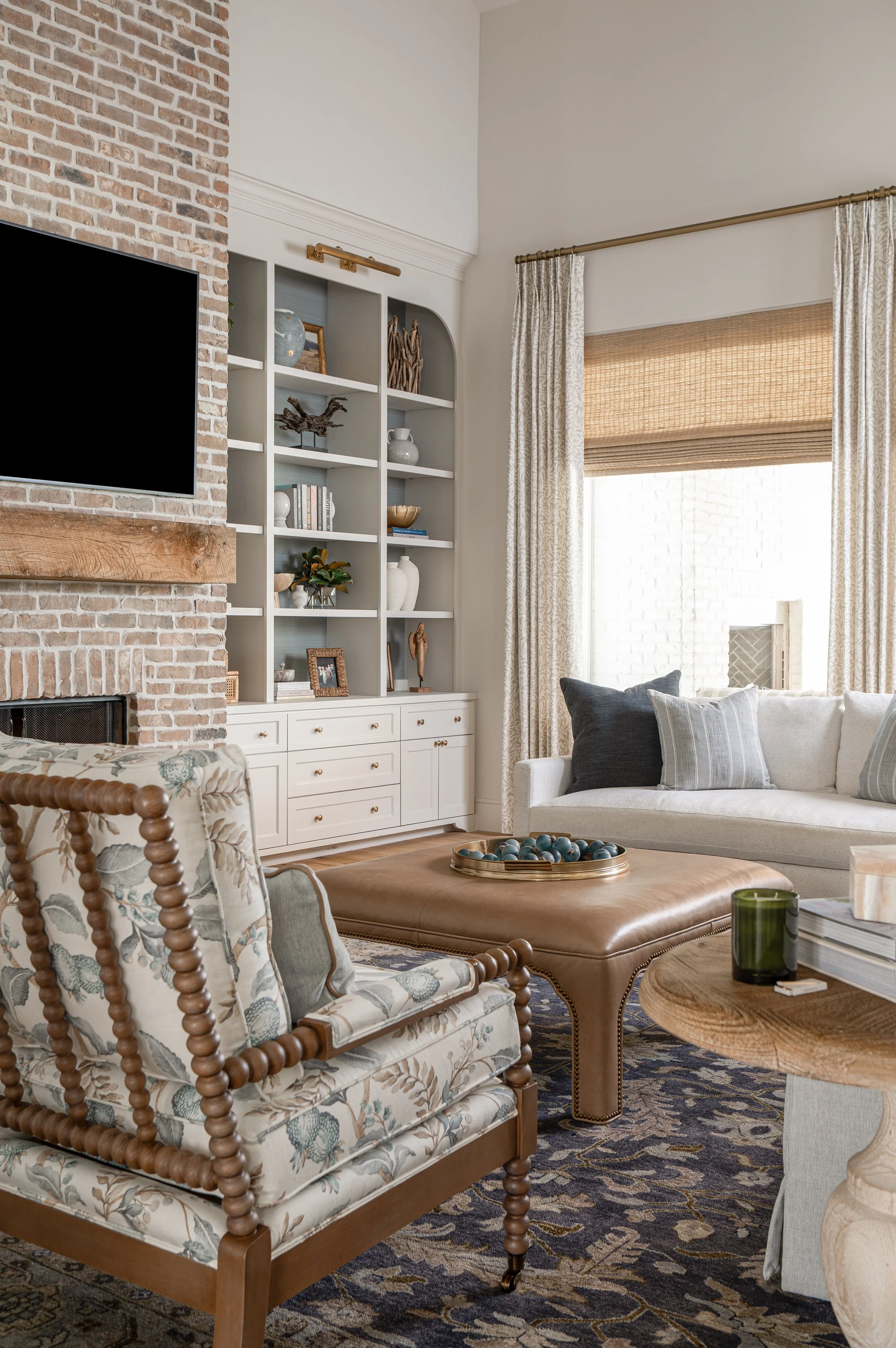 Living room with brick fireplace, built-in white bookshelf with decorative items and books, window with beige shade and floral curtains, beige sofa with pillows, wooden coffee table with tray of decorative balls, patterned area rug.