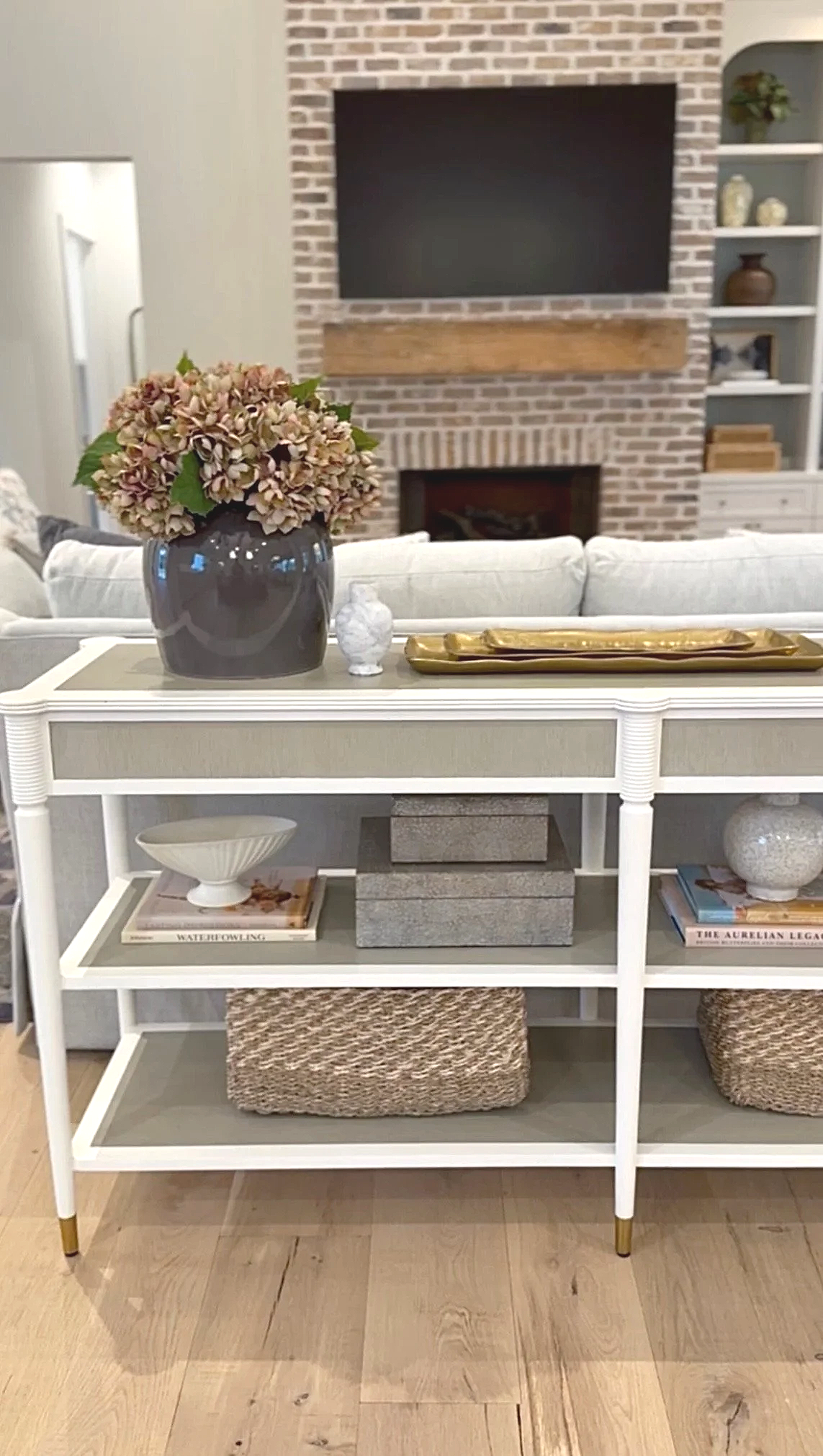 A white console table with decorative items including a dark vase with pinkish flowers, a small white decorative object, and a gold tray. Books and woven baskets are on the lower shelf. In the background, a living room with a brick fireplace and a mounted flat-screen TV are visible.
