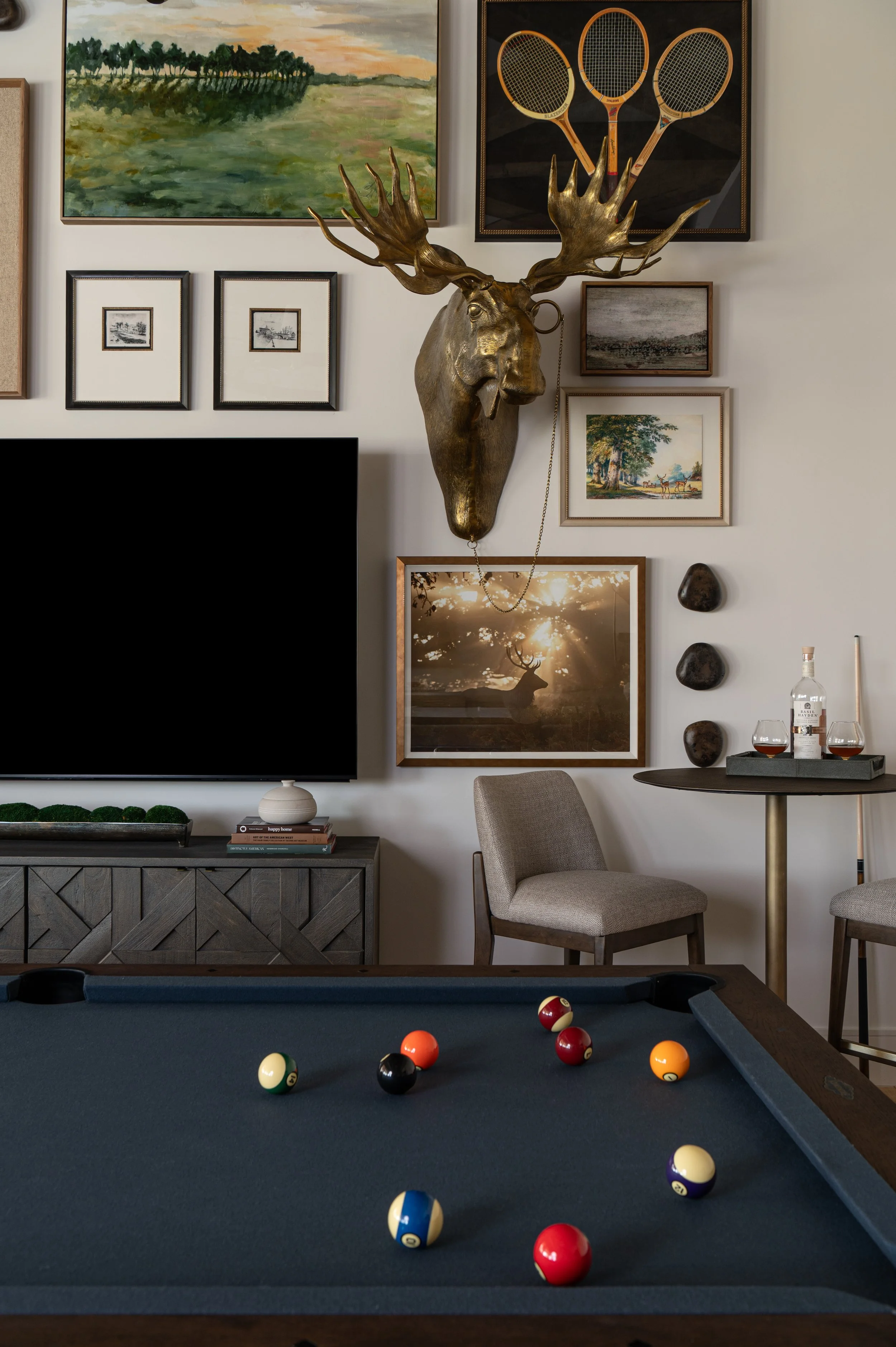 A living room with a pool table, where pool balls are scattered on the table. The background features a gallery wall with framed artwork, a decorative moose head with antlers, and a small round table with glasses and a bottle.