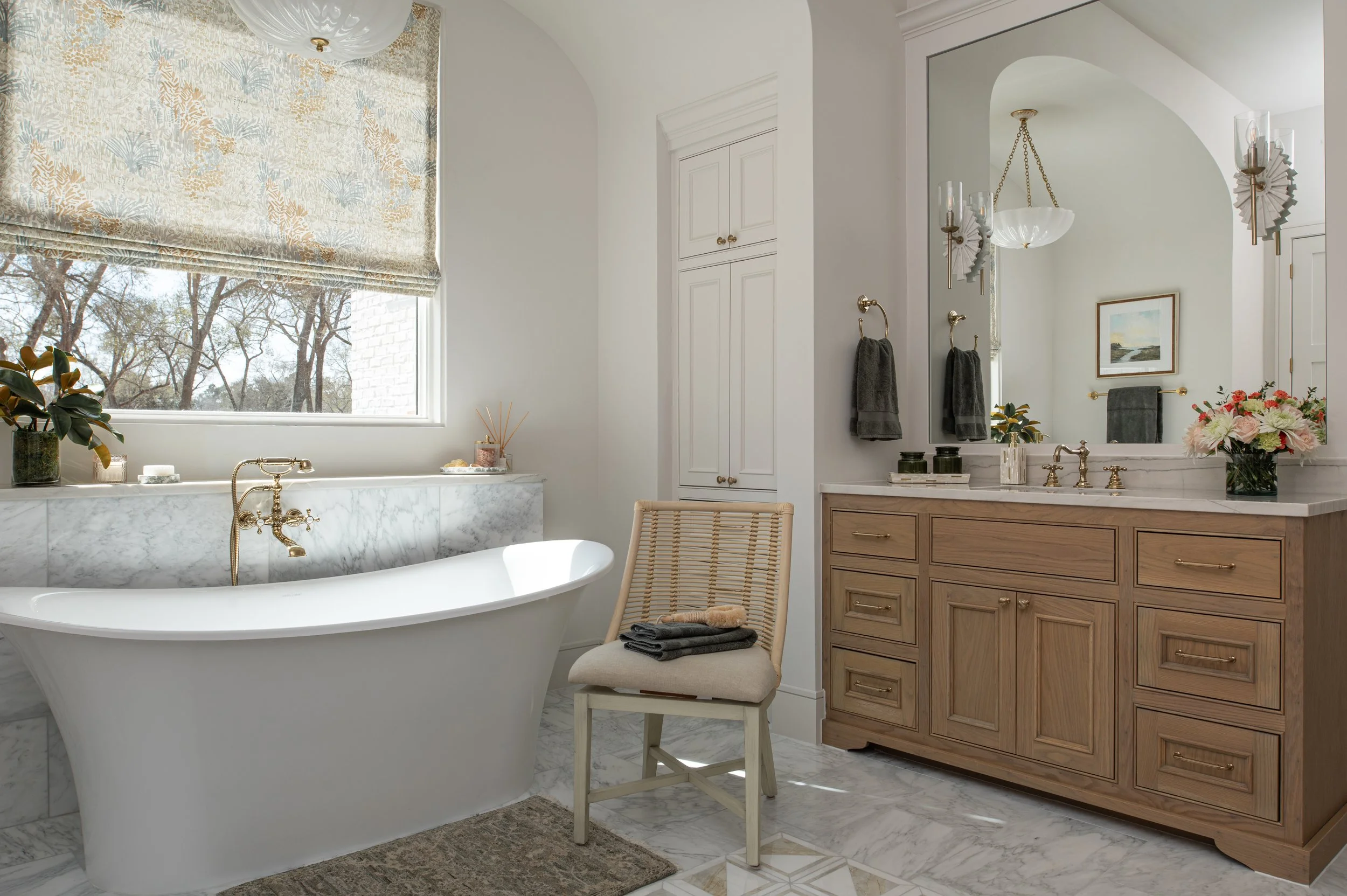 A bright bathroom with a white freestanding bathtub, a large window with a patterned Roman shade, a wooden vanity with framed mirror, and a bouquet of pink flowers on the countertop.