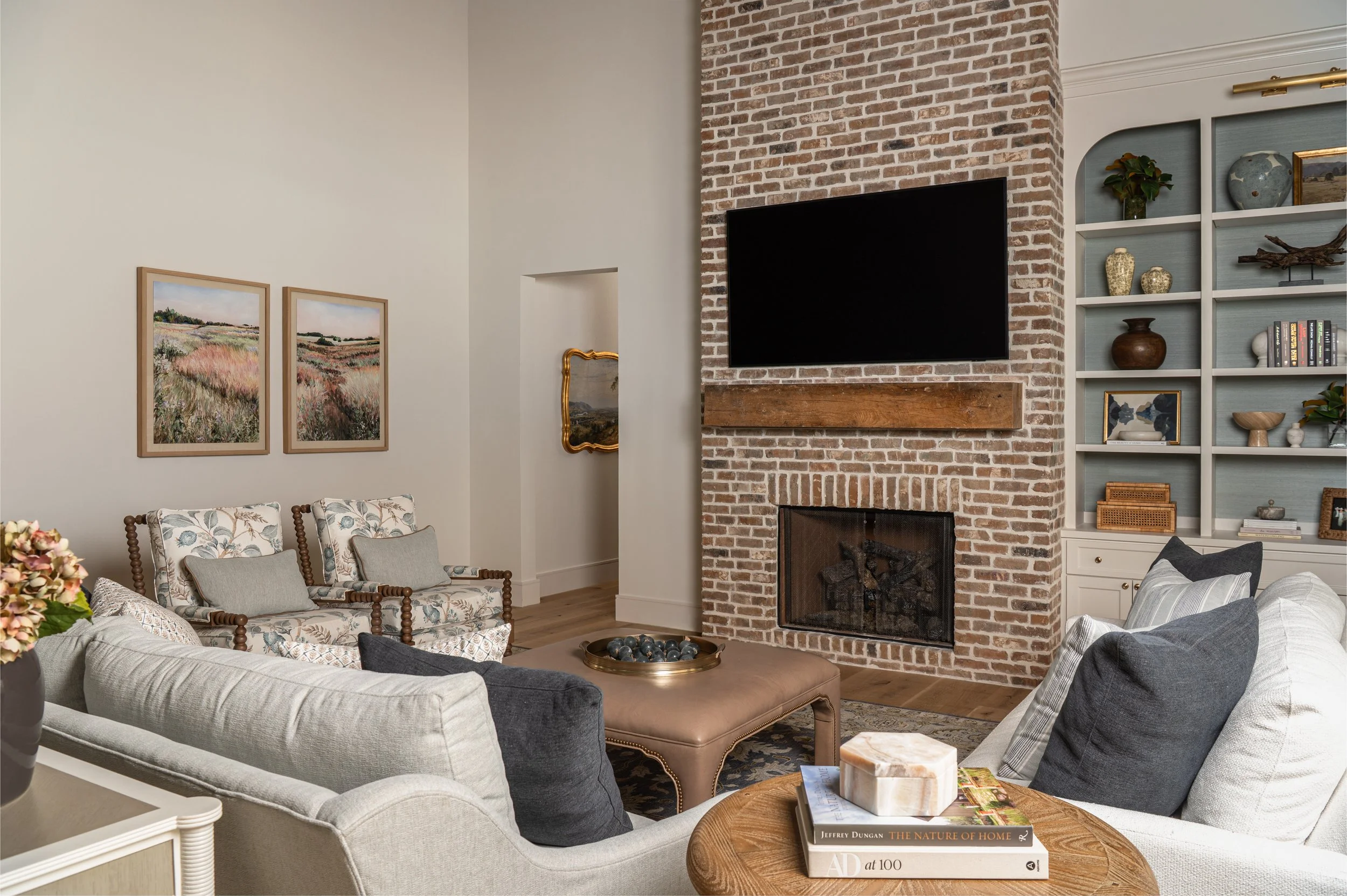 Living room with brick fireplace, mounted flat-screen TV, built-in white bookshelf, and various decorative items. Gray upholstered sofa, floral armchair, wooden coffee table with books, and framed artwork on white walls.
