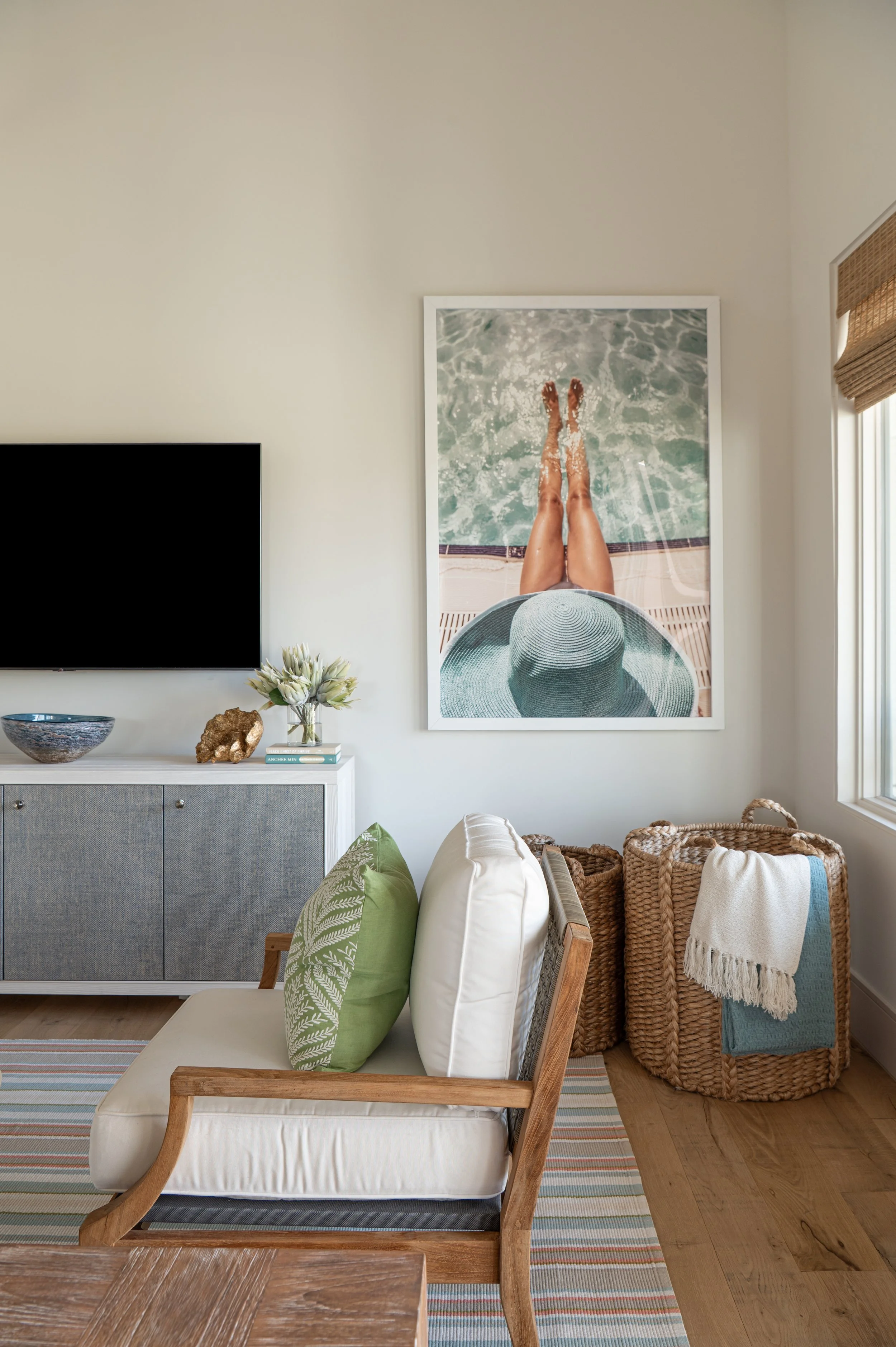 Living room with a white couch, green and white pillows, a wicker basket, a striped rug, a modern TV, a glass vase with flowers, a decorative rock, a bookshelf, and a large framed photo of a person with a sun hat sitting by a pool.