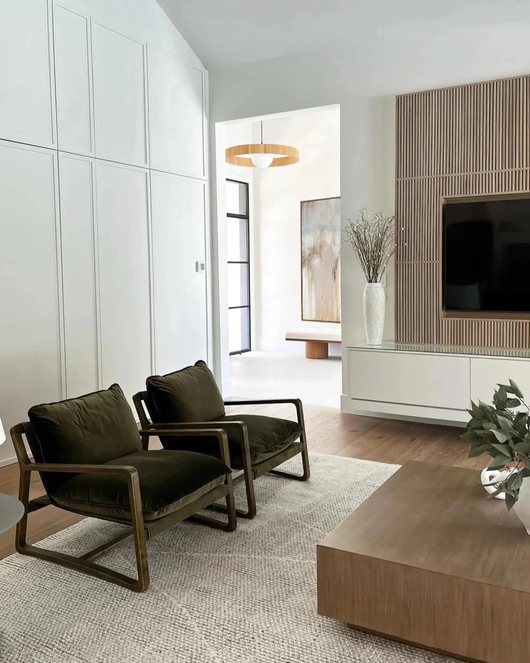 Modern living room with two black cushioned chairs, a beige area rug, a wooden side table, white walls, a large artwork, a white vase with dried plants, and a built-in wooden panel with a television.