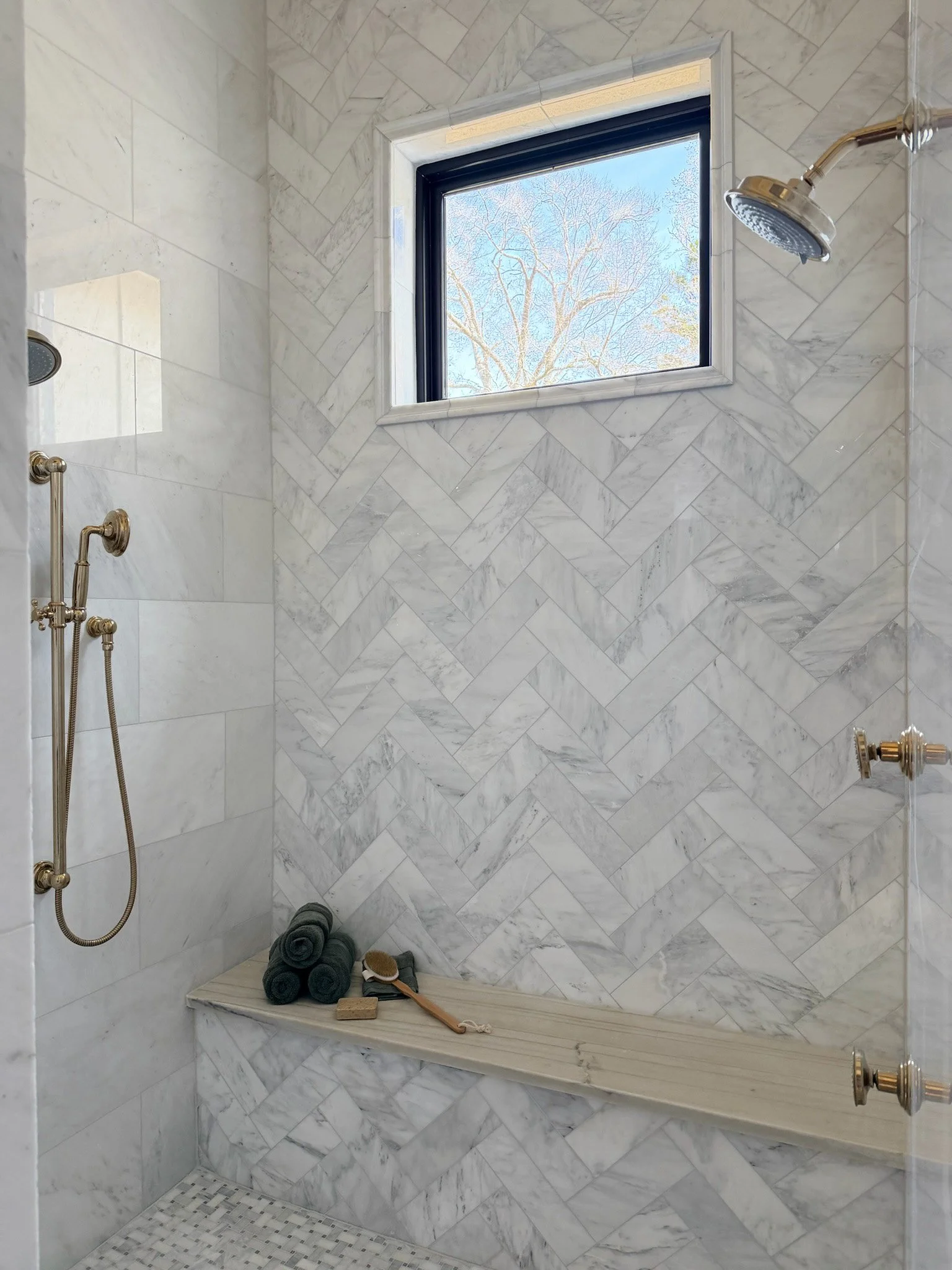 A marble shower with two showerheads, a window showing a tree outside, and a small bench with rolled towels, a brush, and a sponge.
