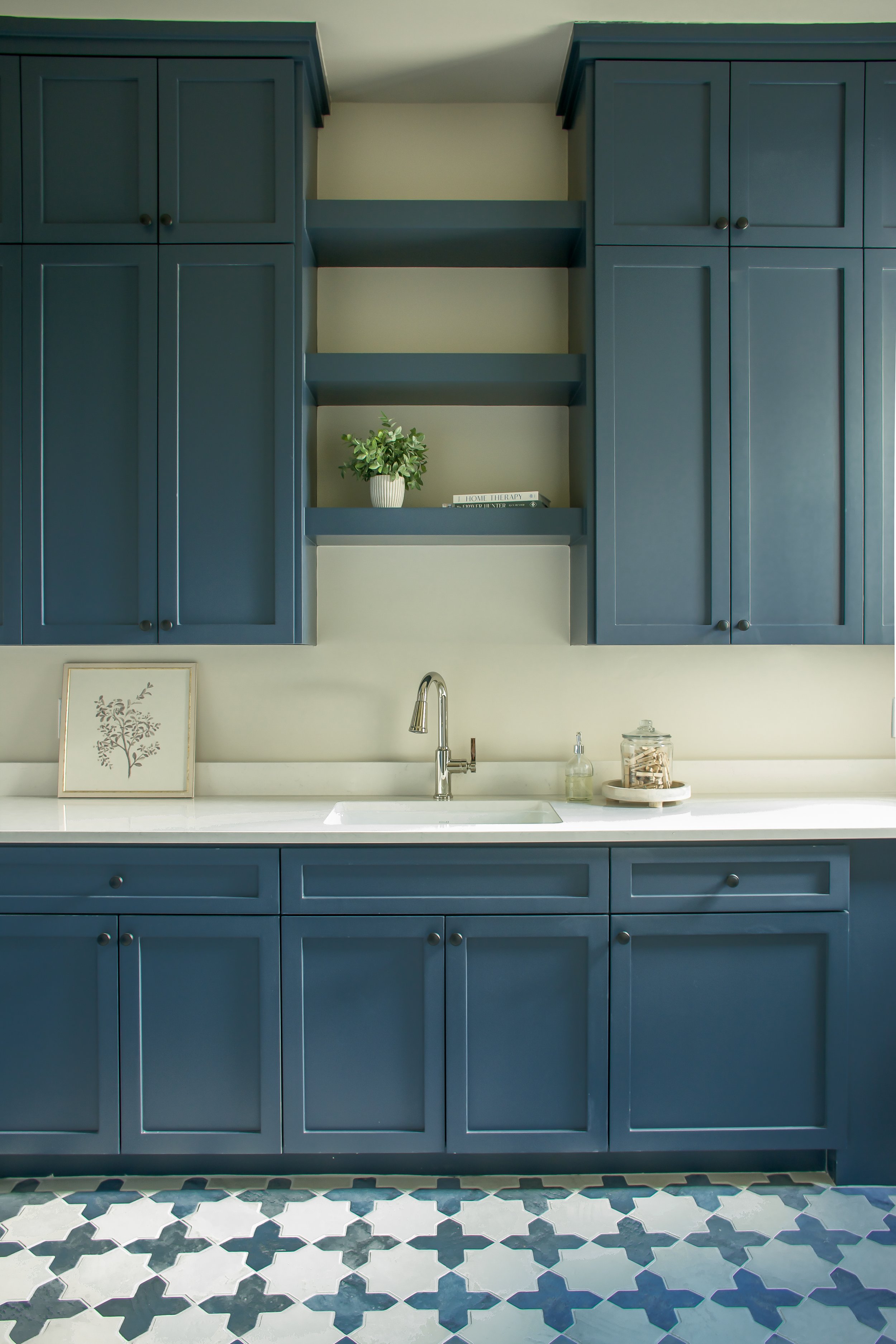 Kitchen with dark blue cabinets, white countertop, a sink with a silver faucet, a small decorative framed picture of a tree, a glass jar, and a small tray with a jar of cotton swabs. The lower part of the wall is wallpapered with a blue and white geo