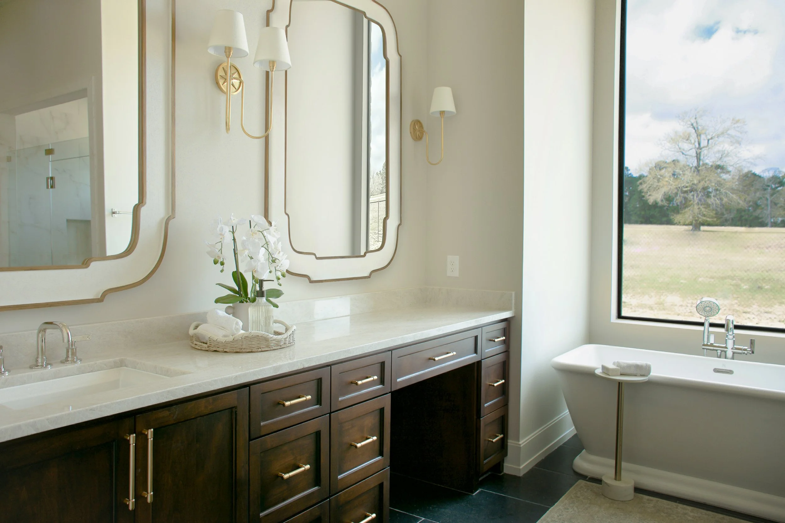 A modern bathroom with a white marble countertop, dark wood cabinets, and a large window showing a tree outside. There are two large mirrors with decorative gold framing, wall-mounted white lamps, a orchid plant in a glass vase, and a bathtub with a 