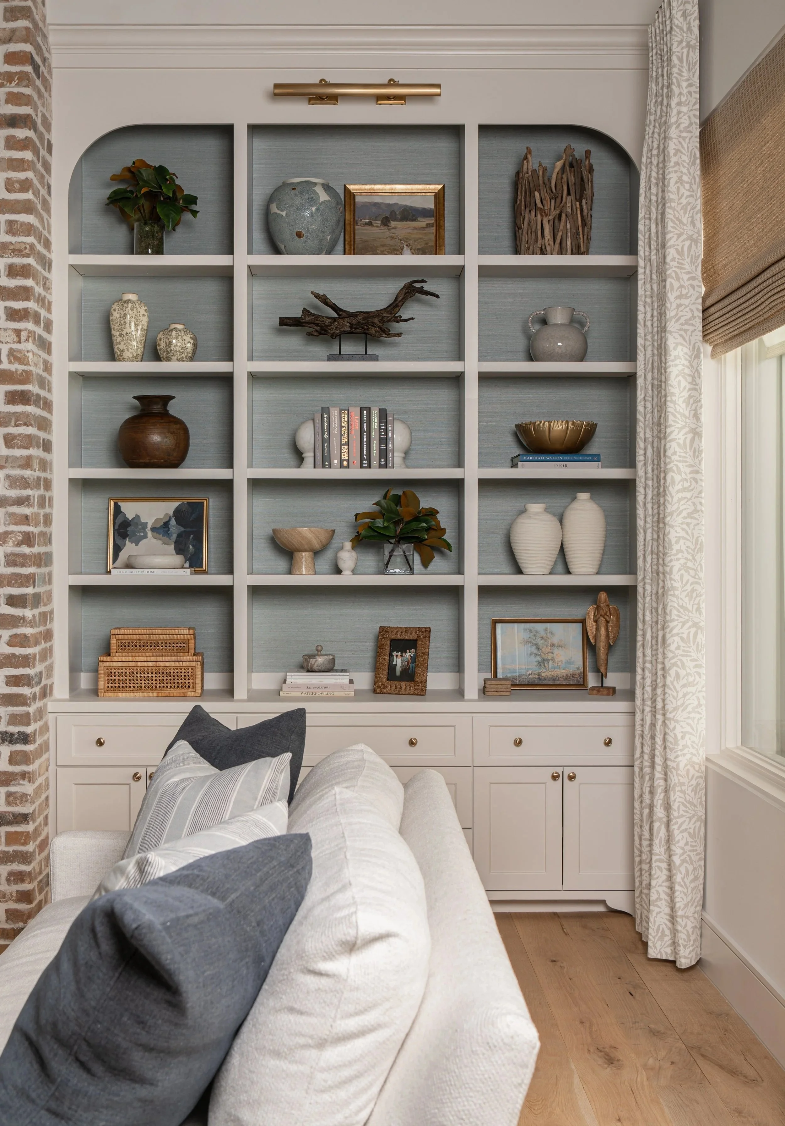 Living room corner with a beige sofa, striped pillows, white built-in bookshelf with various decorative items like vases, artwork, driftwood, books, and framed photos, adjacent to a brick wall and window with patterned curtains.