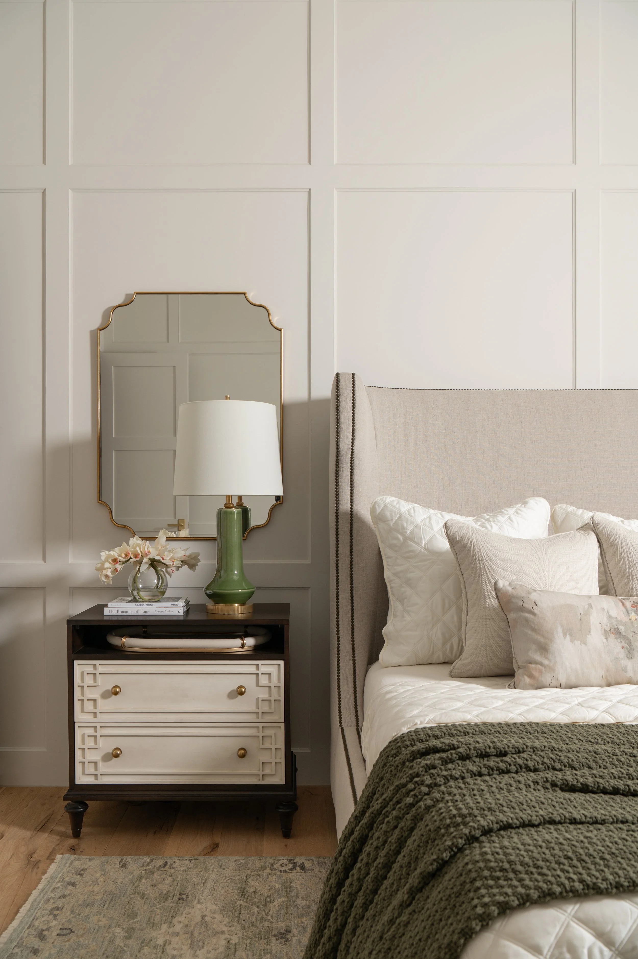 A bedroom with a beige upholstered bed, beige pillows, a dark side table with a green lamp, a mirror with a gold frame, and a vase of pink flowers. The wall behind has decorative white paneling.