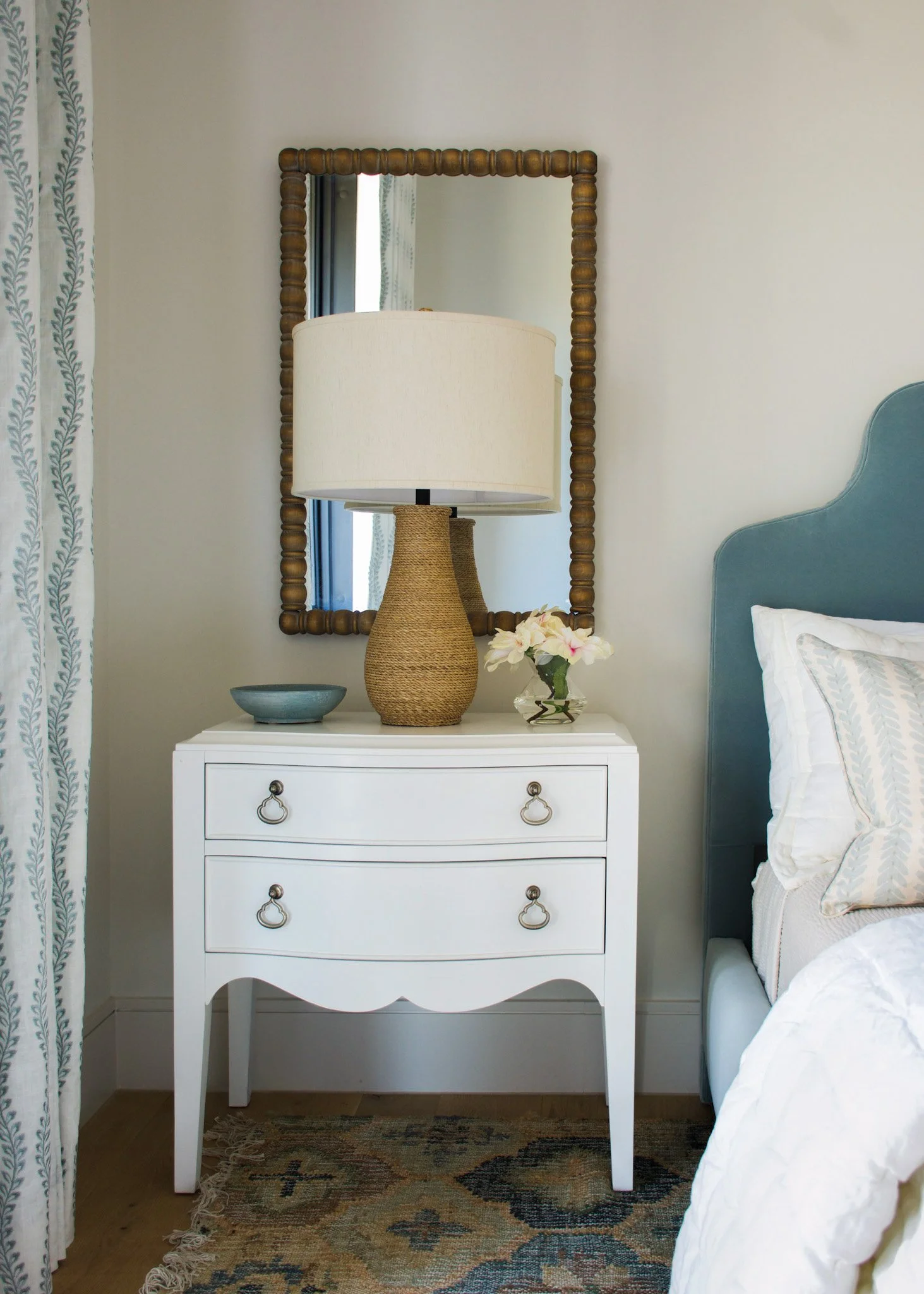 A white bedside table with three drawers and metal ring pulls, topped with a beige woven lamp, a small glass vase with pink and white flowers, and a blue ceramic bowl. Behind the table, a rectangular wooden mirror hangs on the wall, reflecting a wind