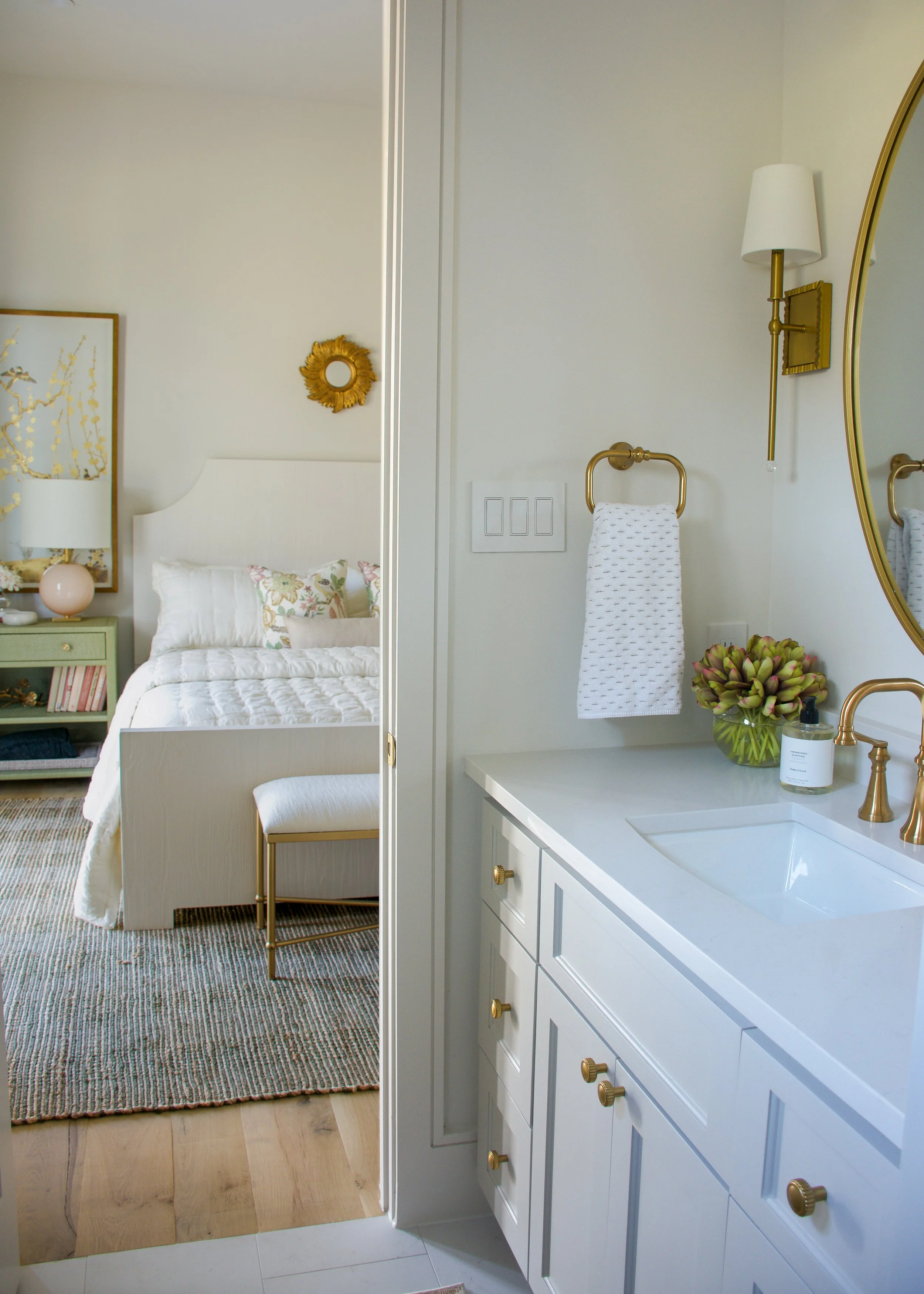 A bedroom visible through a doorway, with a white bed with floral pillows, a green nightstand with a pink lamp, and a wall with artwork and a golden mirror. The foreground shows a white bathroom vanity with a gold faucet, a white countertop with a so