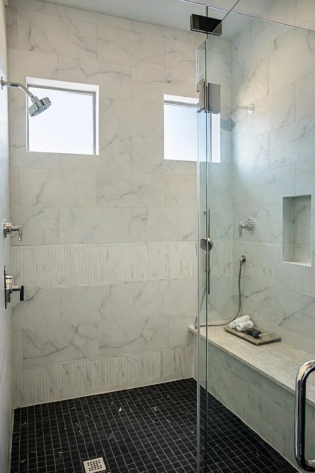 Modern shower with marble walls, two small windows, a rainfall showerhead, and a built-in bench with rolled towels.