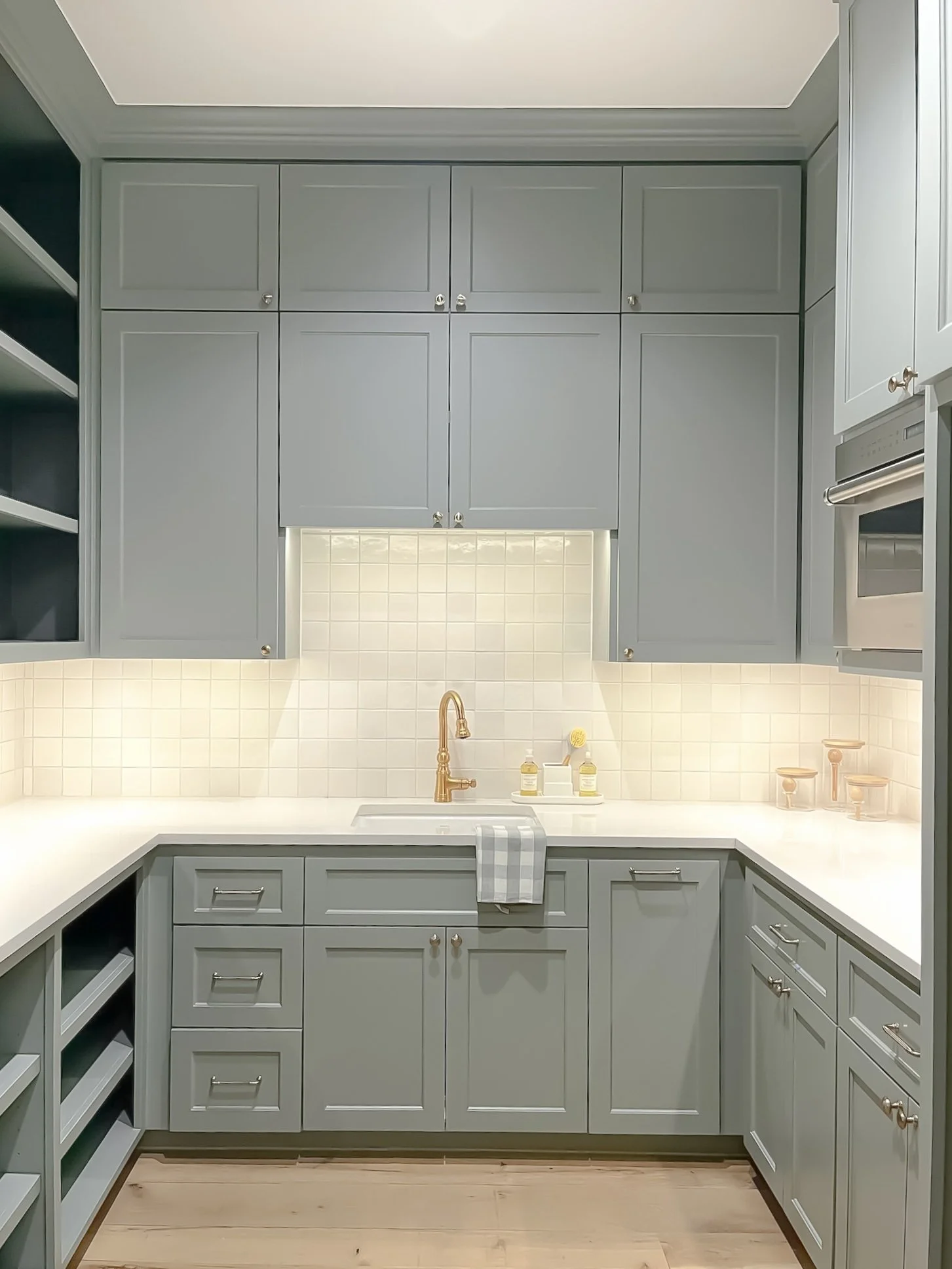 A kitchen with light gray cabinets, a white countertop, a gold faucet, and a white tiled backsplash. Open shelving on the left, some bottles and glasses on the counter, and a dish towel hanging over the sink.