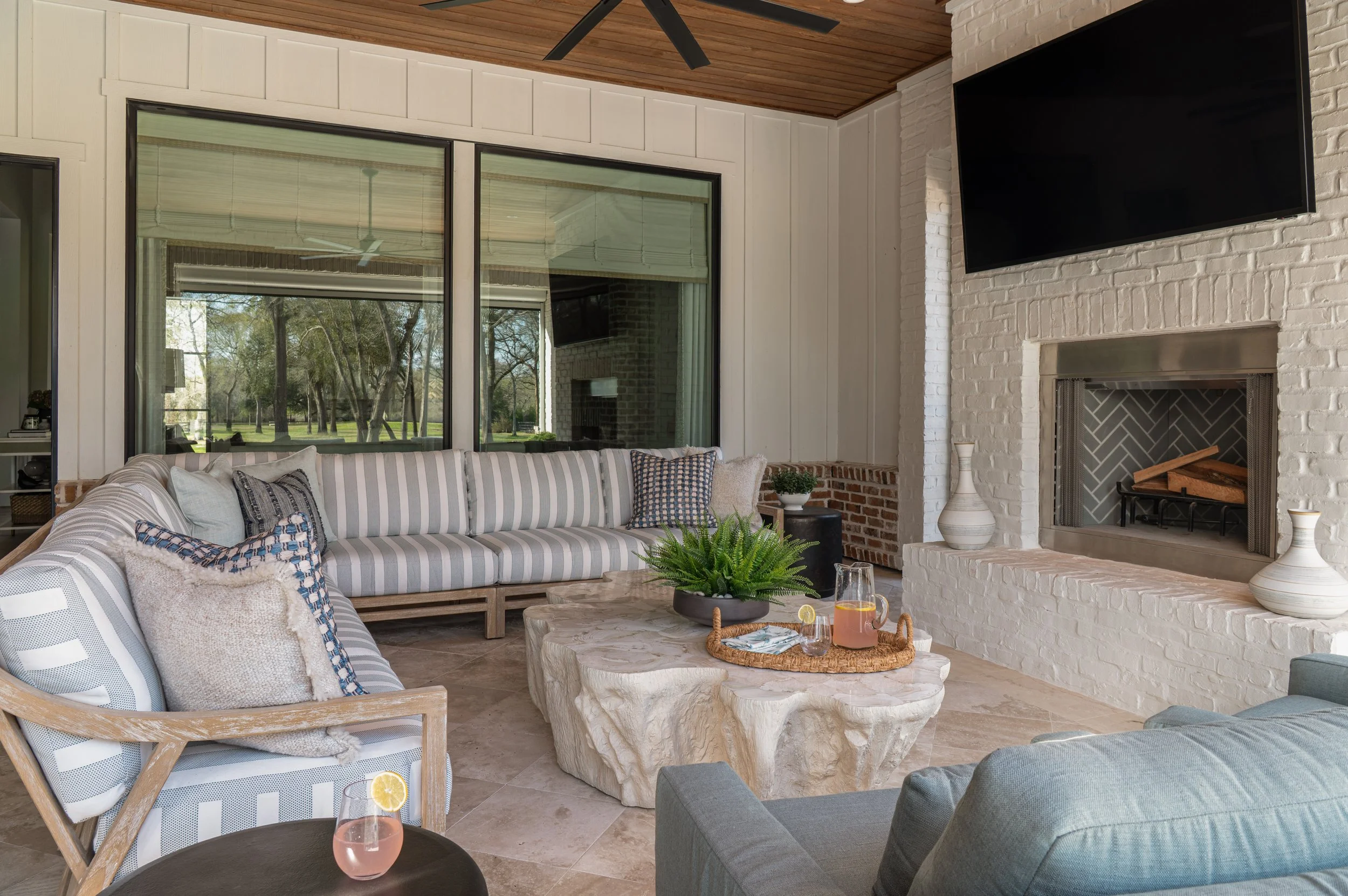 Living room with large windows showing trees outside, featuring a gray and white striped couch, round marble coffee table with decorative vases, a black side table with a plant, a fireplace with decorative vases, a mounted TV, and a wood-paneled ceil