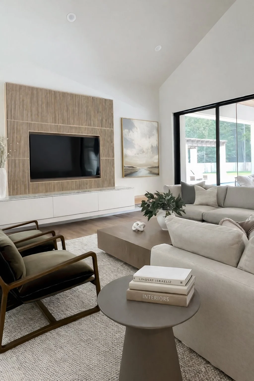 Modern living room with beige sectional sofa, black-framed sliding glass door, wall-mounted flat-screen TV, wooden accent wall, abstract landscape painting, coffee table with plant and books, armchair, and a textured area rug.
