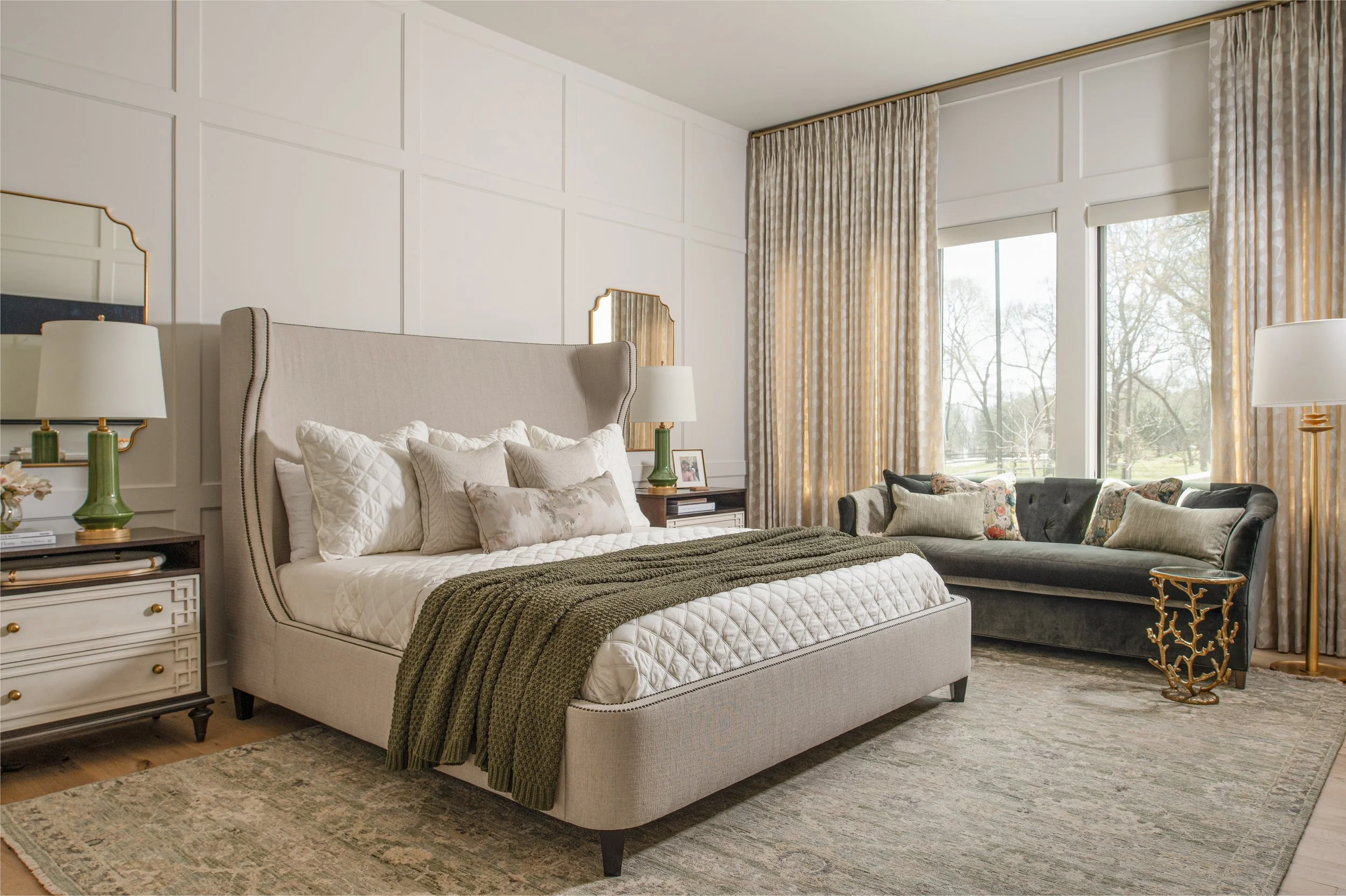 A bedroom with a beige upholstered bed, nightstands with green lamps, a dark gray sofa with patterned pillows, tall windows with beige curtains, and a large area rug.