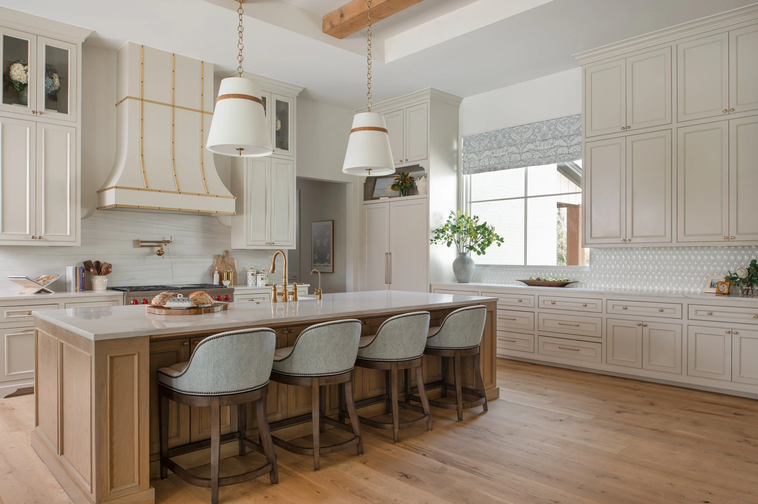 A modern kitchen with white cabinets, a large island with a wood base, upholstered chairs, a window with a blue patterned Roman shade, and decorated with plants and kitchenware.