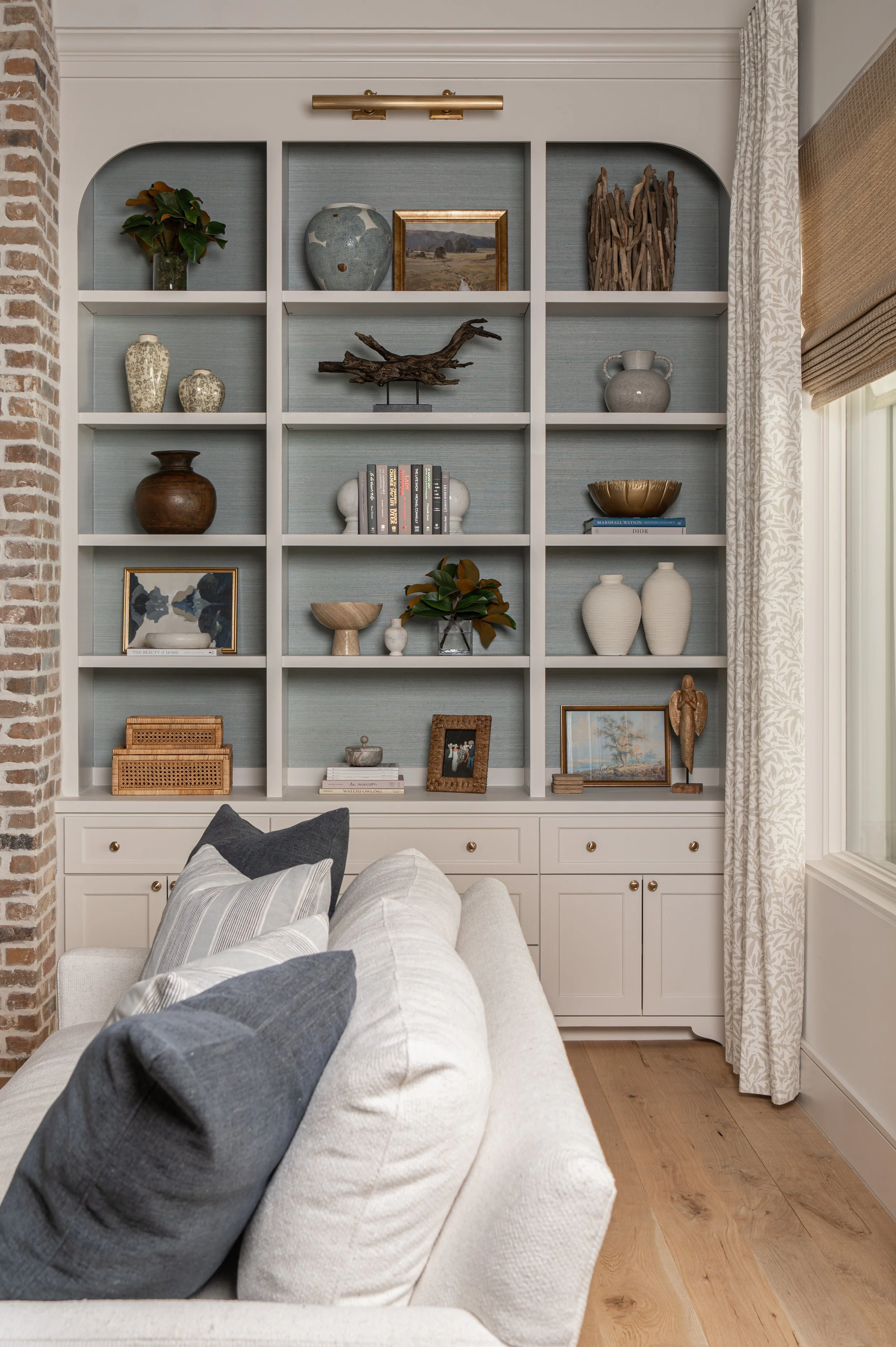 A living room with a built-in white bookshelf filled with various decorative items, books, and artwork. In front of the bookshelf, there is a light-colored sofa with striped and solid-colored pillows. To the right, there are curtains and a large wind